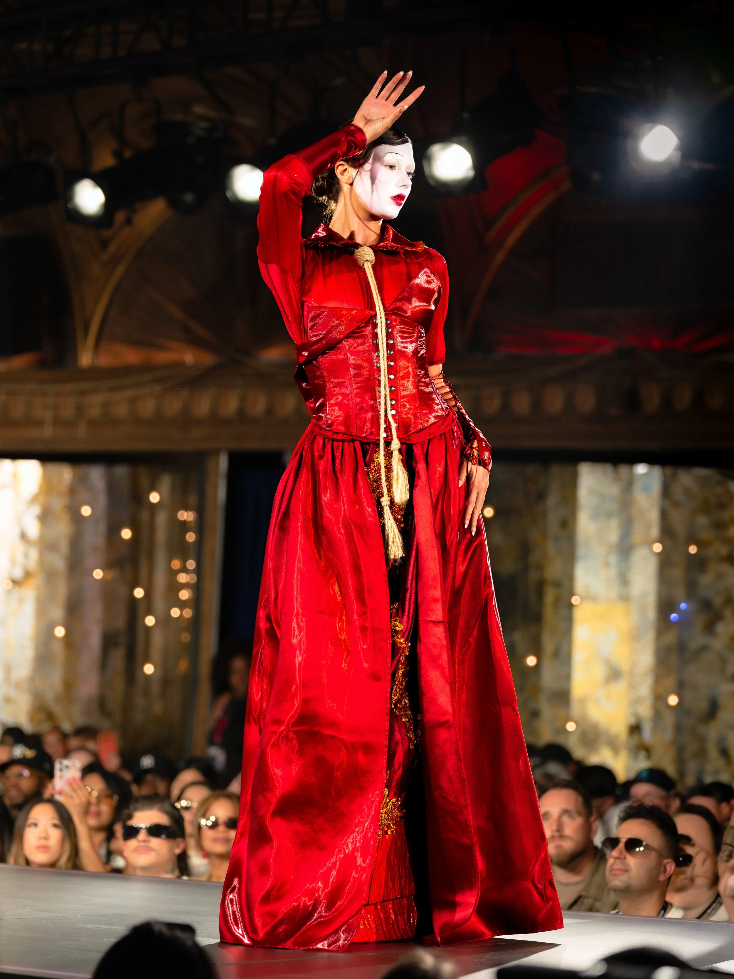 A woman with pale face makeup, dark hair, red lipstick, and dressed in a red satin dress with gold embroidery, walking on a runway at a fashion show. She's raising her right hand and looks elegant and poised. The background shows an ornate, classical