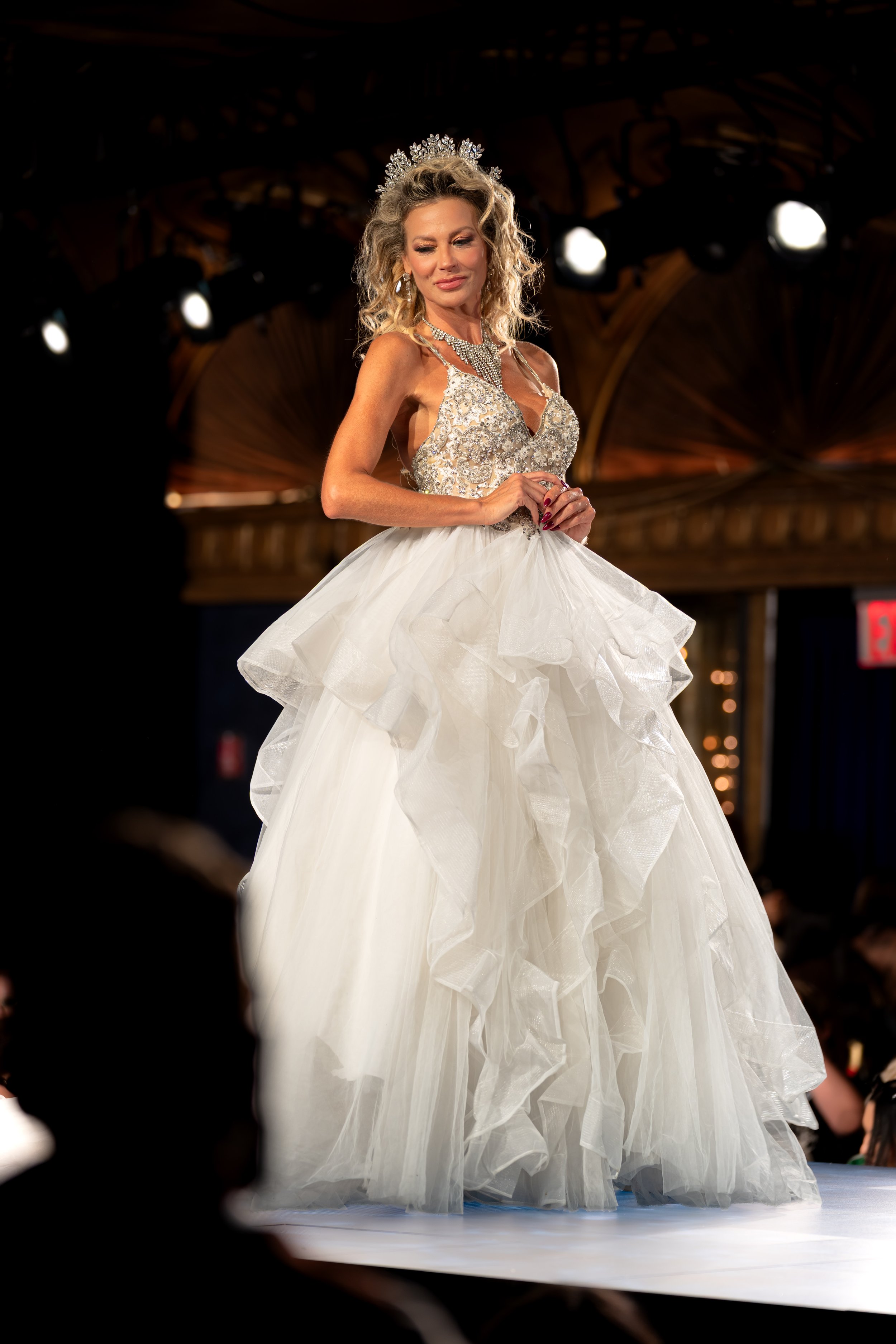 A woman in a wedding gown on a runway, wearing a tiara and jewelry, with a dark, ornate background and audience in the distance.