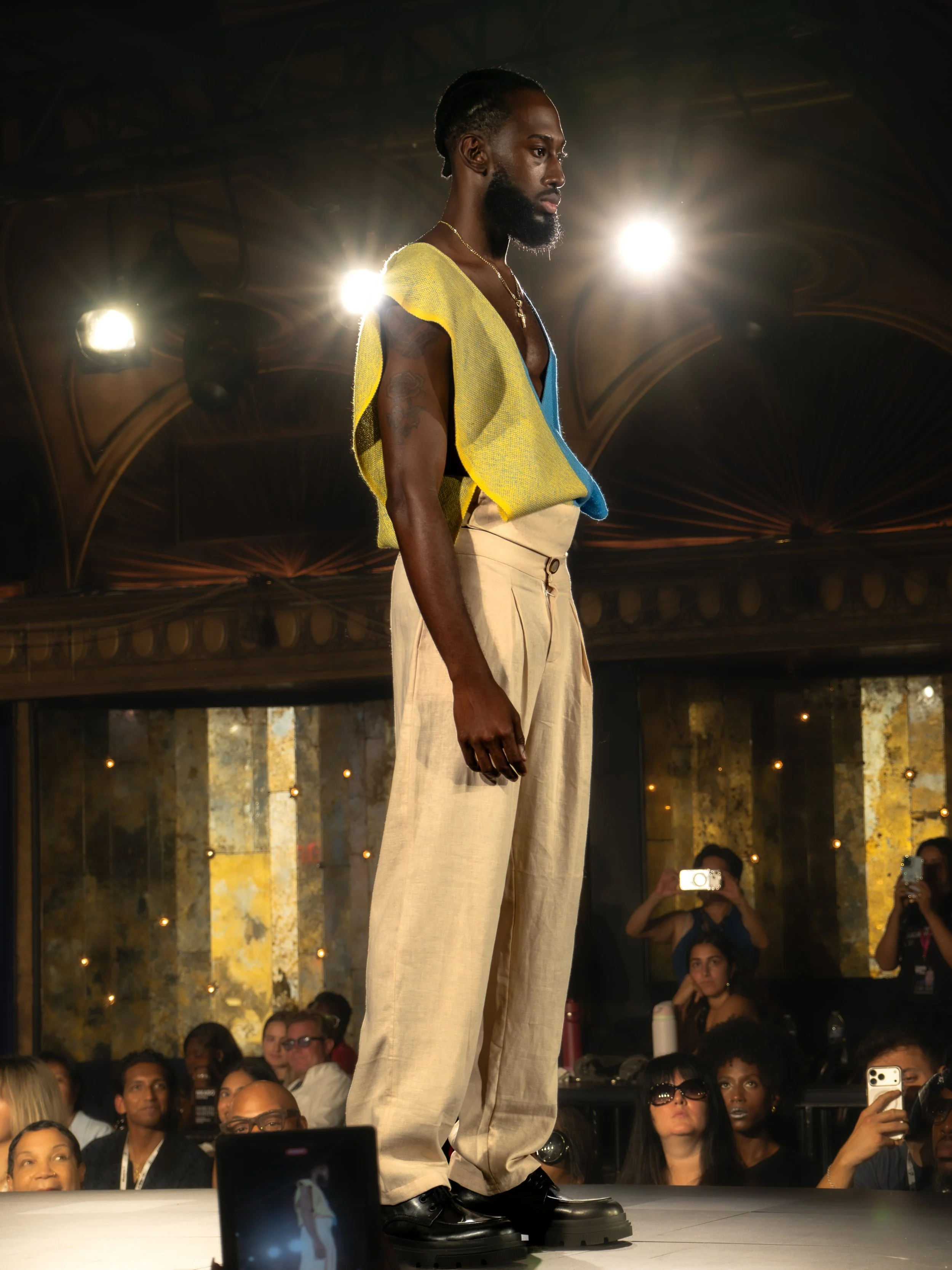 A man with dark skin and a beard wearing a yellow and blue vest, beige wide-leg pants, and black boots standing on a runway during a fashion show, with an audience watching in the background and stage lights behind him.