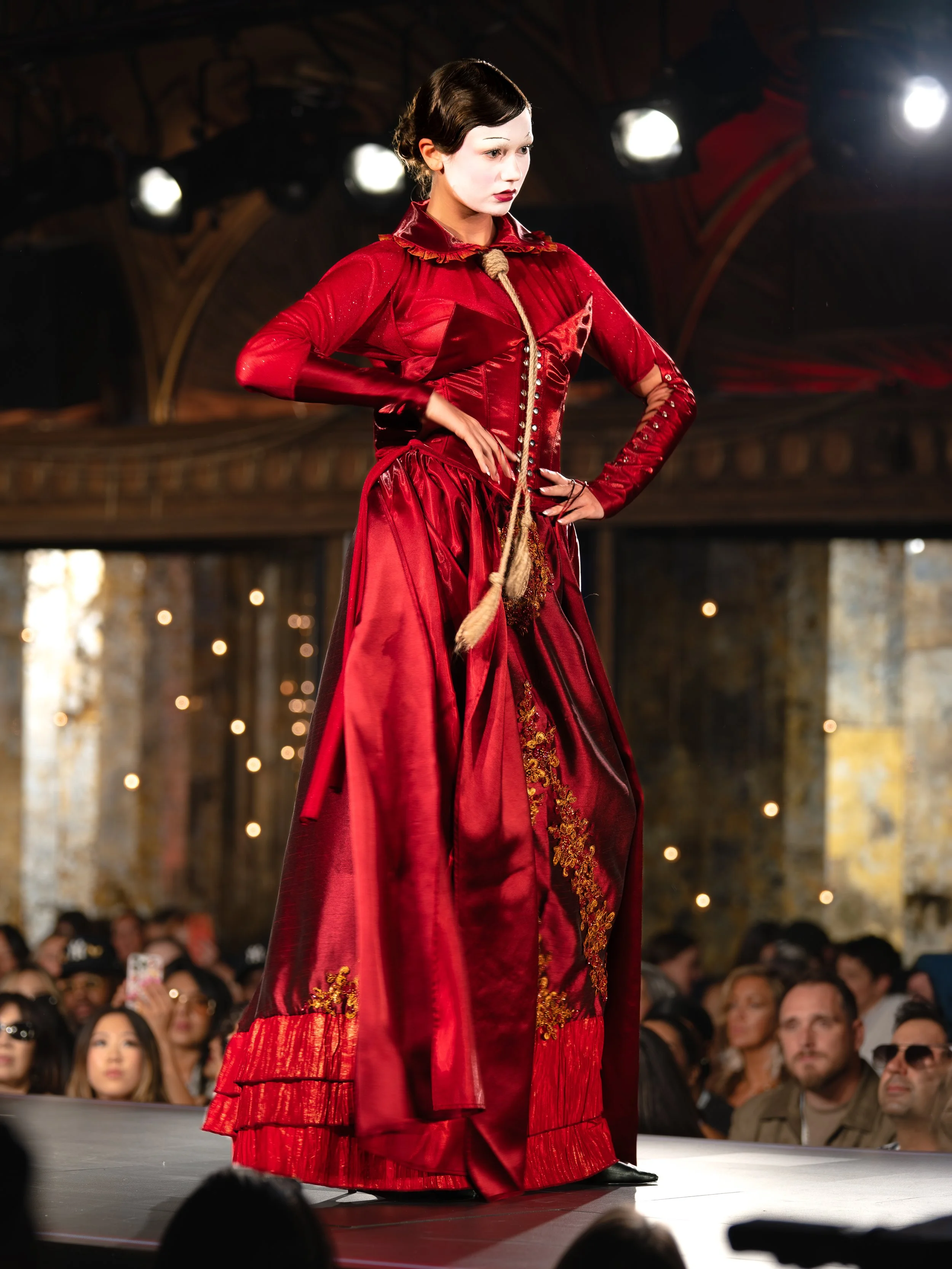 A model on a runway wearing a long, elegant red dress with gold embroidery and fringe details, surrounded by an audience and stage lights.