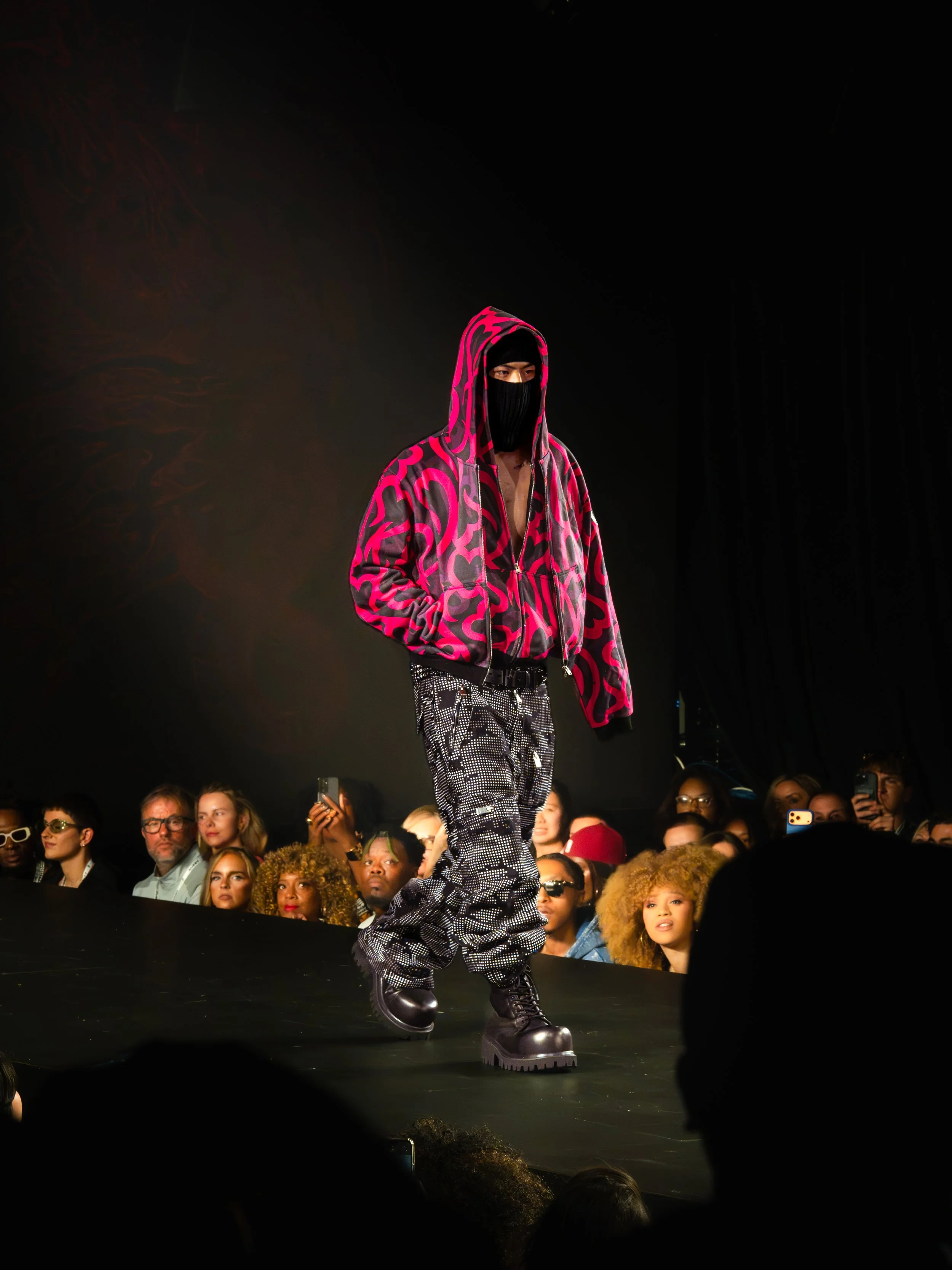 Fashion model walking on runway wearing a pink and black patterned hoodie, black face mask, black platform boots, and black and white patterned pants, with an audience watching.