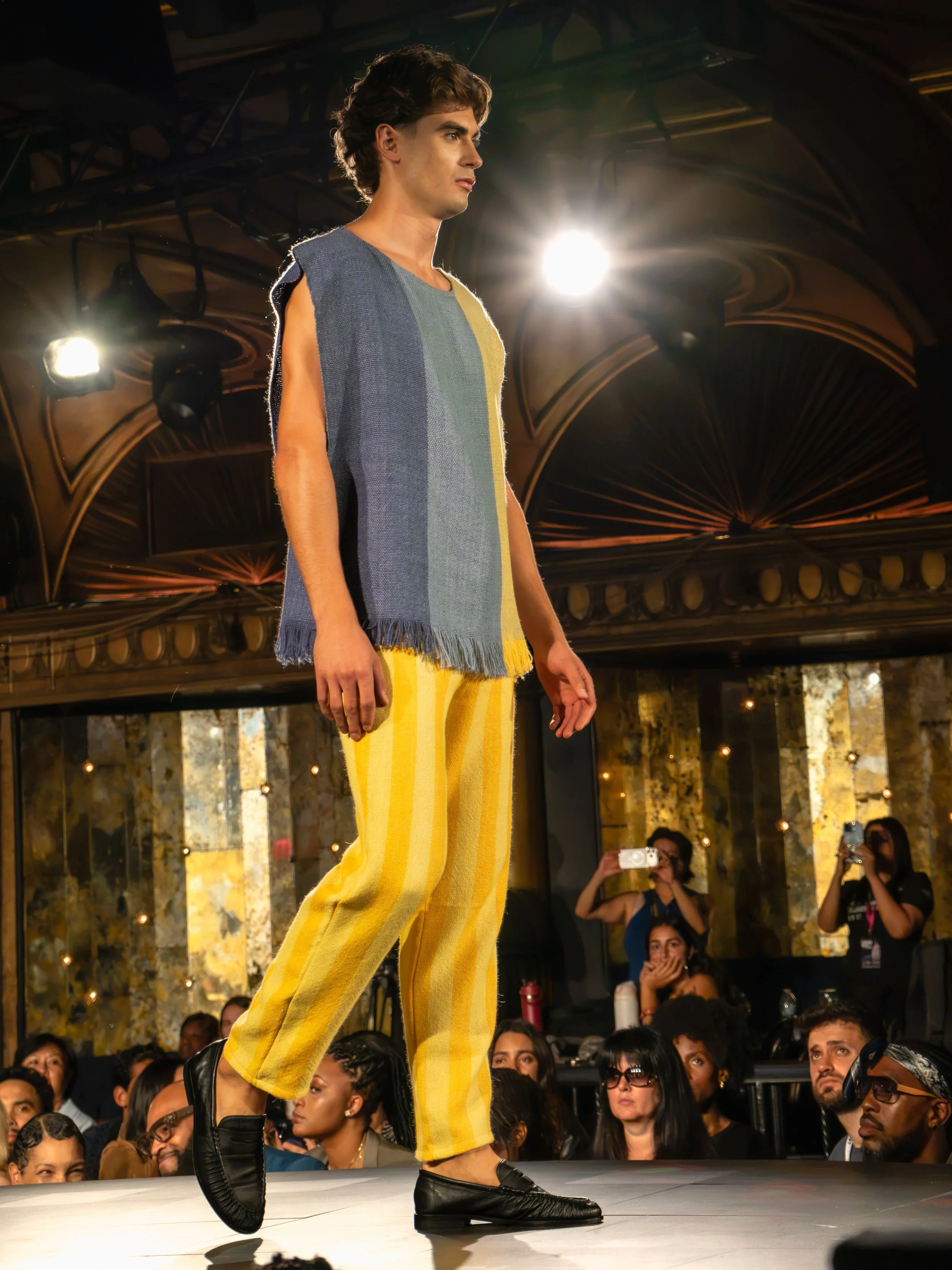 A male model on a runway during a fashion show, wearing a loose, sleeveless, multicolored top and yellow striped wide-leg pants. The model is walking with his right foot lifted, and there are bright stage lights overhead. Audience members and photogr