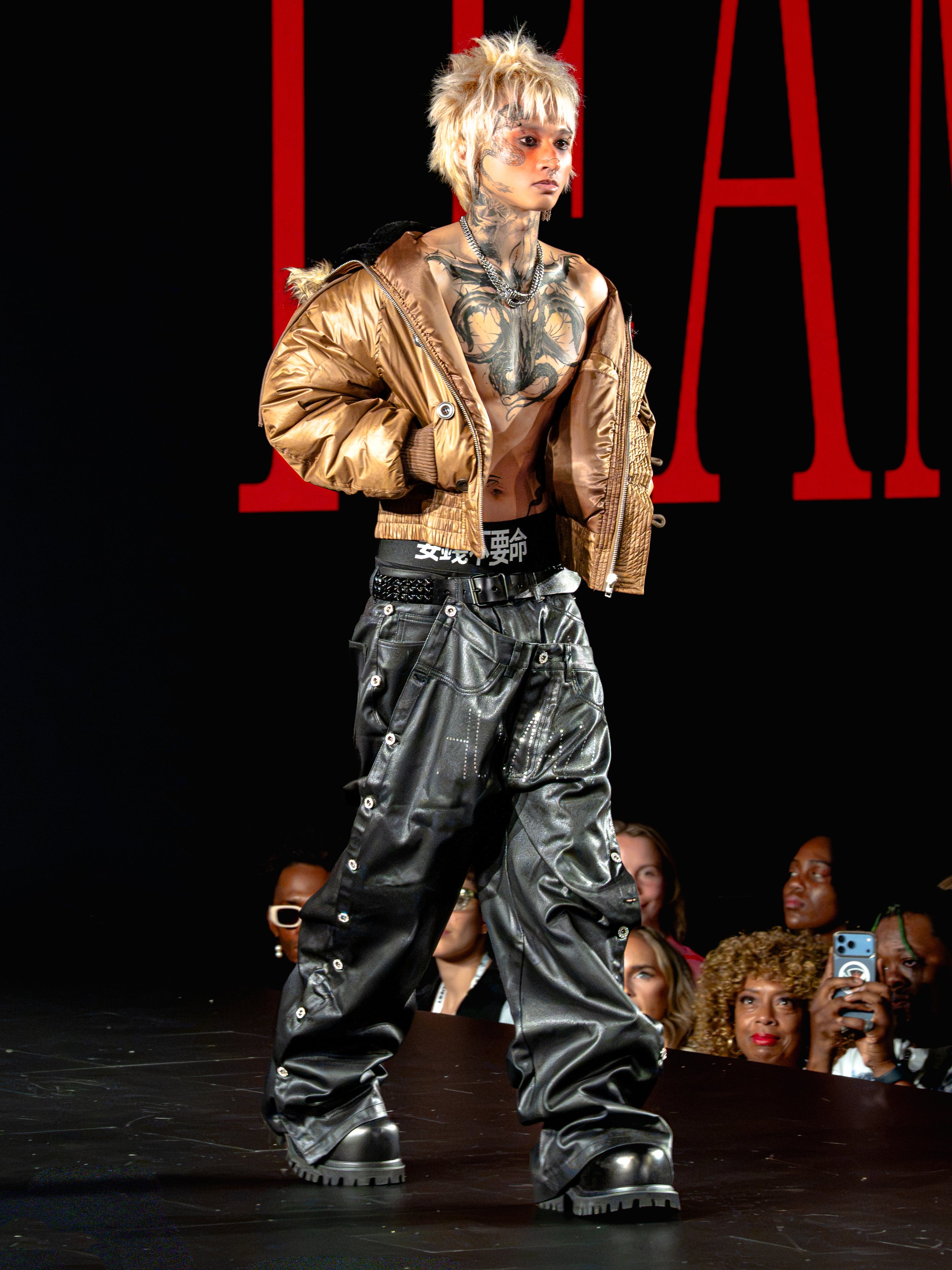 Person with bleached blonde hair, tattoos, wearing a brown jacket, black baggy pants with eyelets, and chunky boots, walking on a runway during a fashion show with an audience in the background.