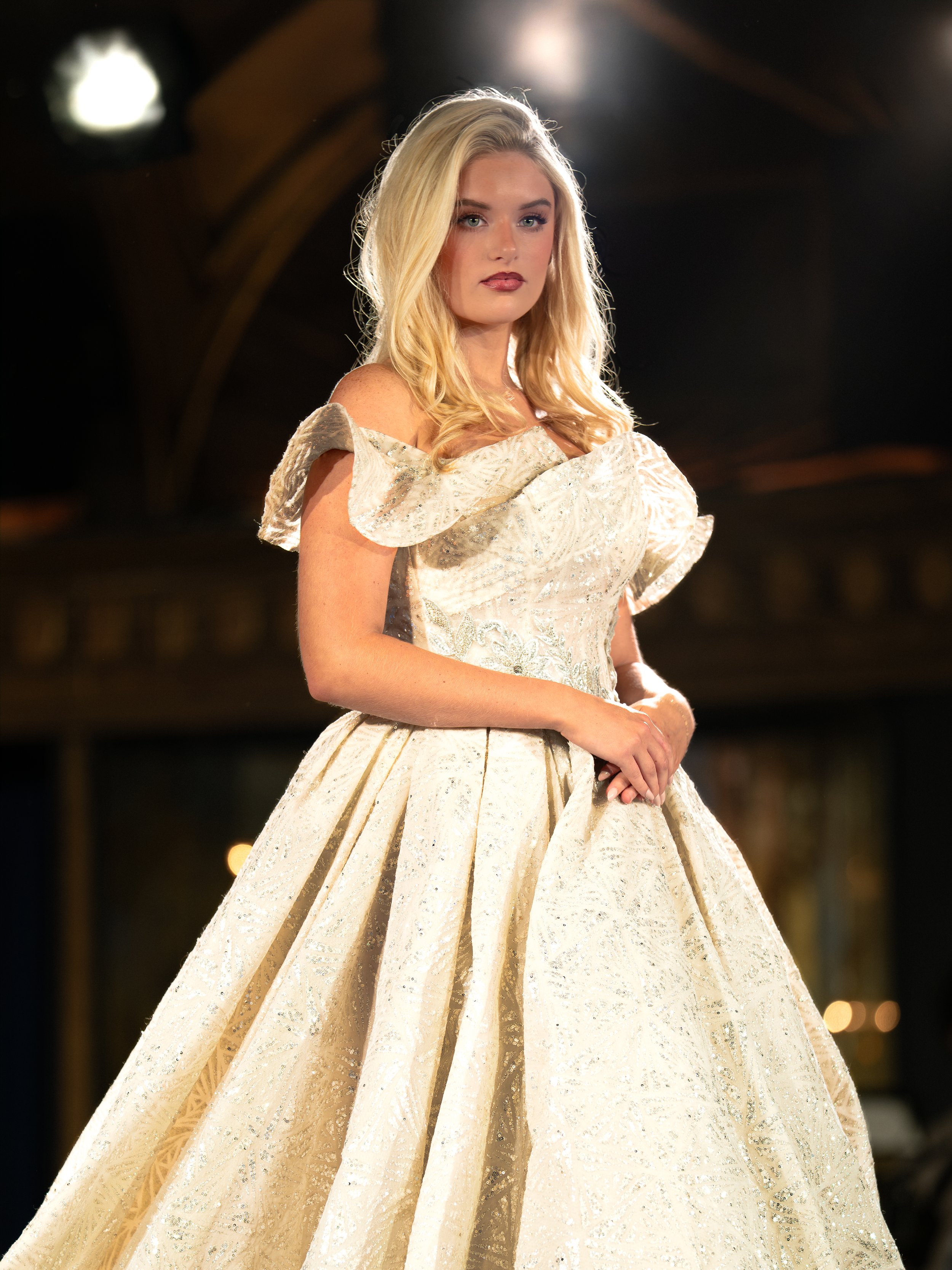 A woman with long blonde hair wears a luxurious, cream-colored ball gown with detailed patterns and ruffles on the shoulders, standing in a dimly lit setting.