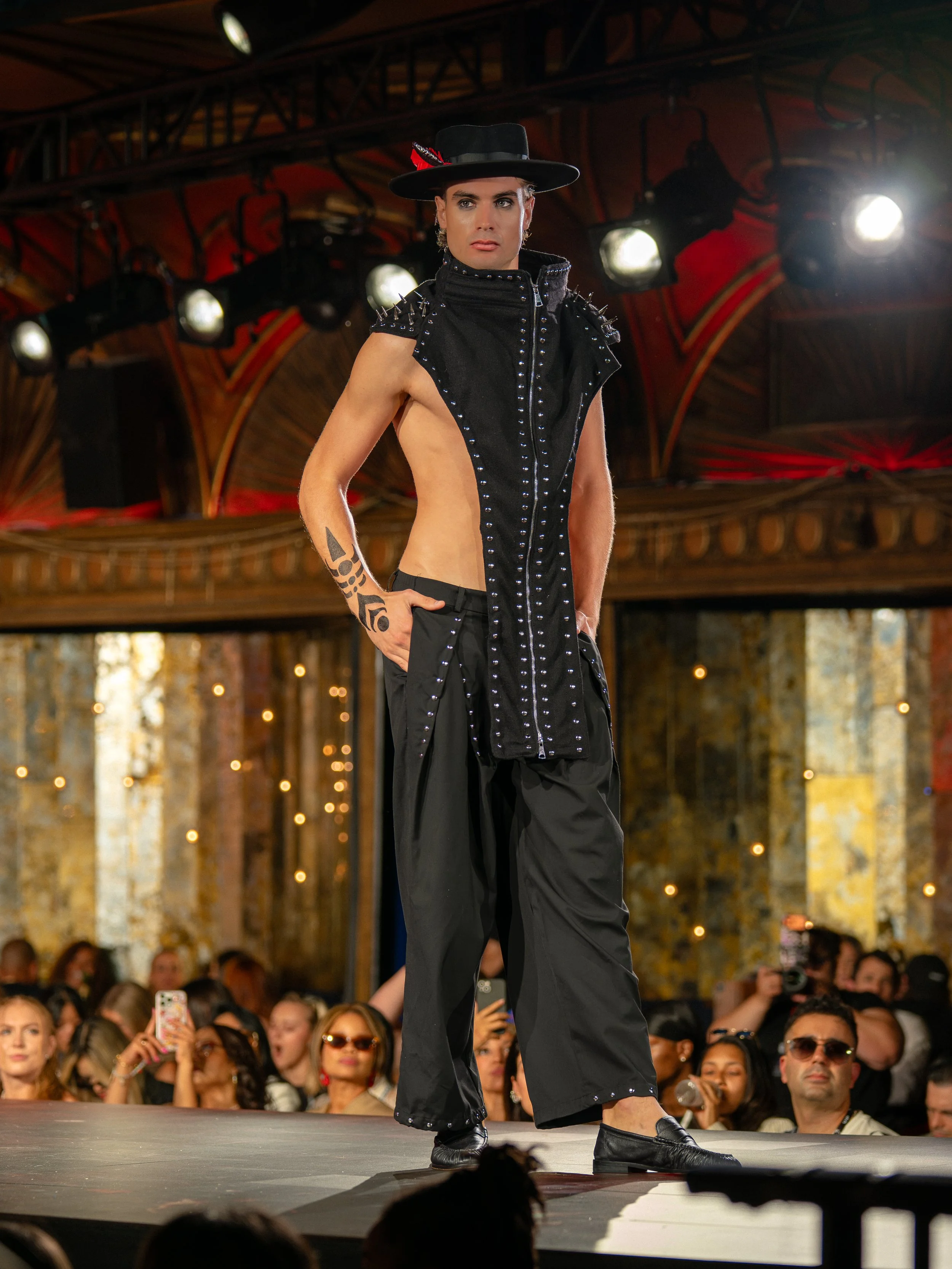 A person dressed in black, wearing a wide-brimmed black hat with a red feather, black sleeveless top with silver studs, large black pants, and black shoes, walking on a runway during a fashion show, with a large audience watching and taking photos in