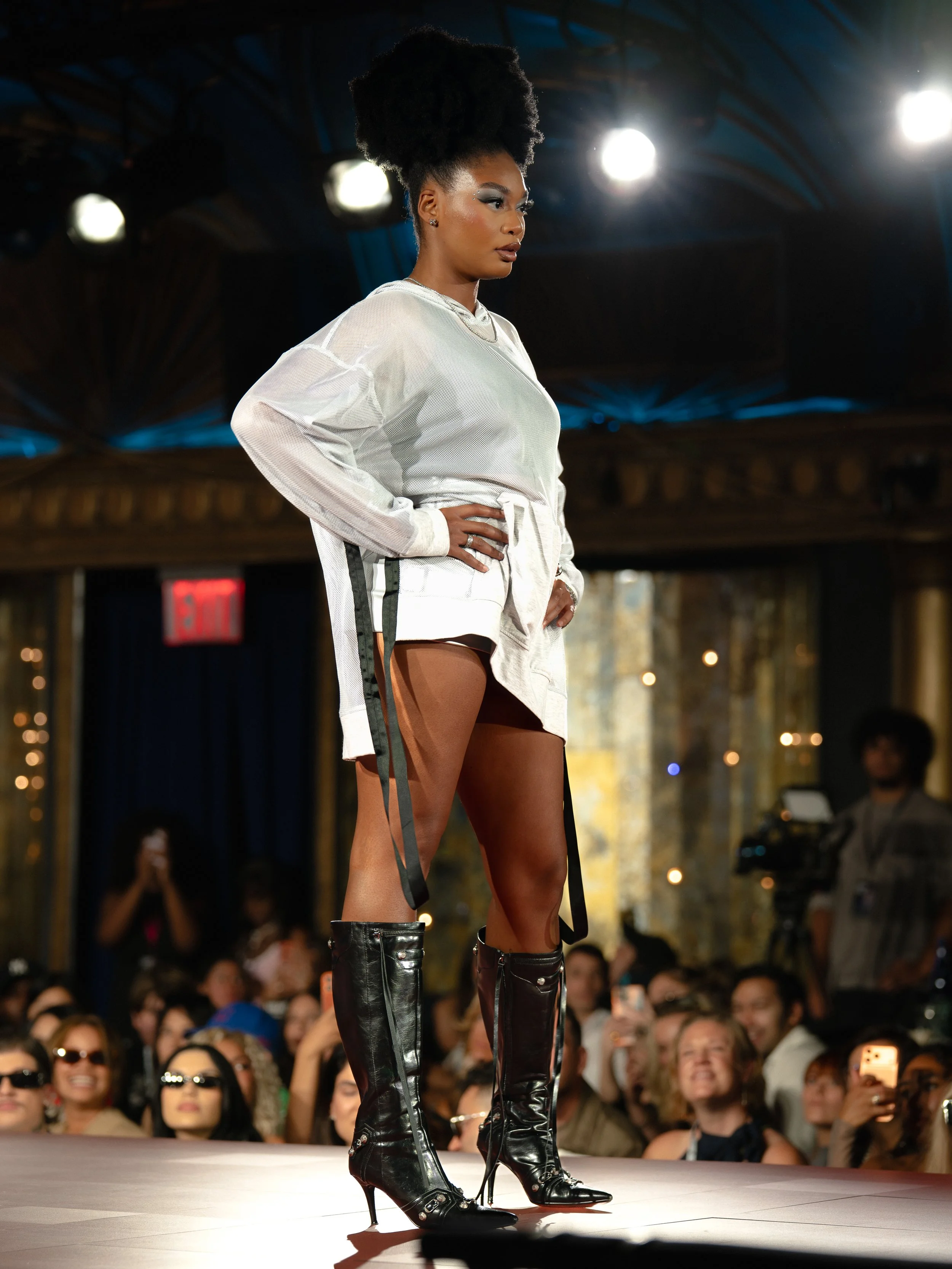 A fashion model walking on a runway during a fashion show, wearing a white outfit with thigh-high black leather boots, and surrounded by an audience taking photos.