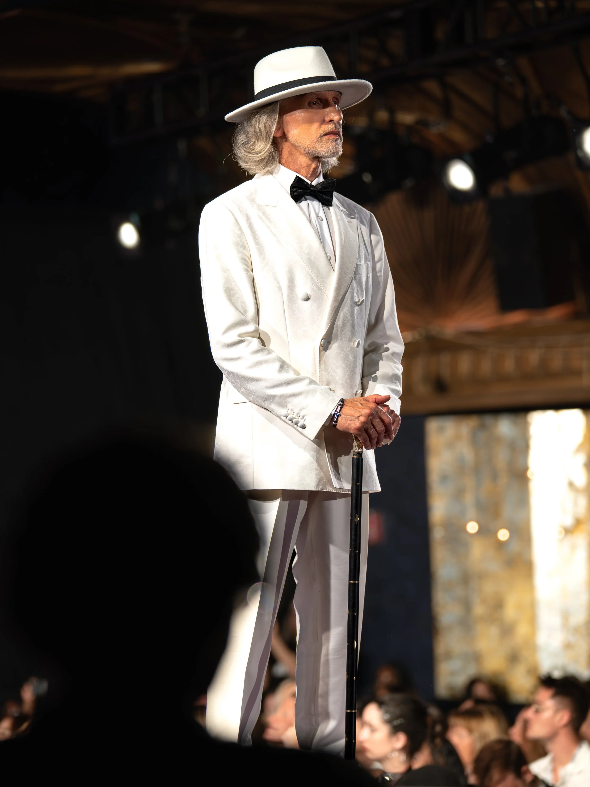 An older man with gray hair and beard, dressed in a white suit with a textured pattern, black bow tie, white hat with black band, and holding a cane, stands on stage at a fashion show or event.
