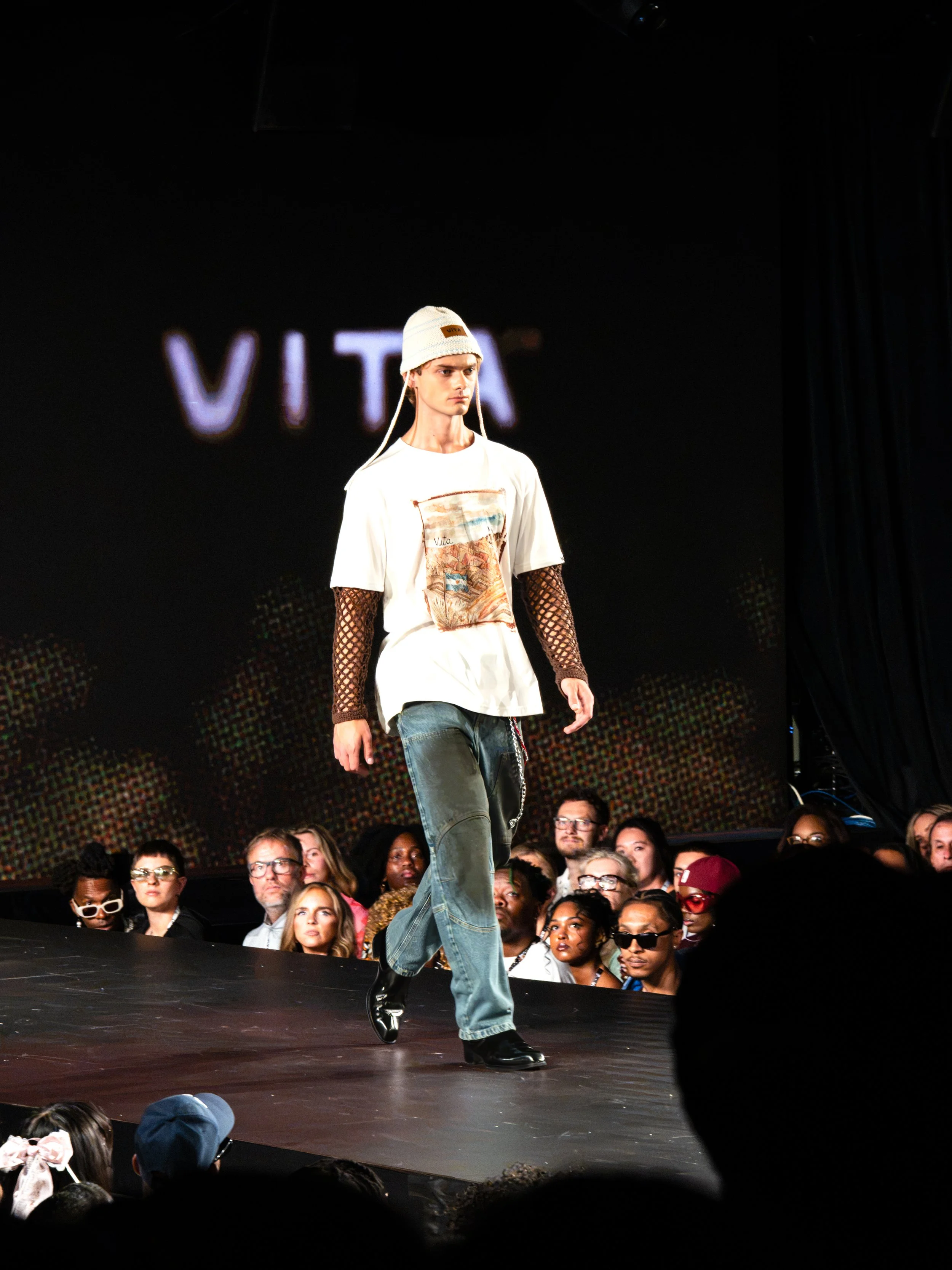 Male runway model walking on stage during a fashion show, wearing a white graphic T-shirt, fishnet sleeves, baggy jeans, black boots, and a white beanie, with a large audience seated around the runway.