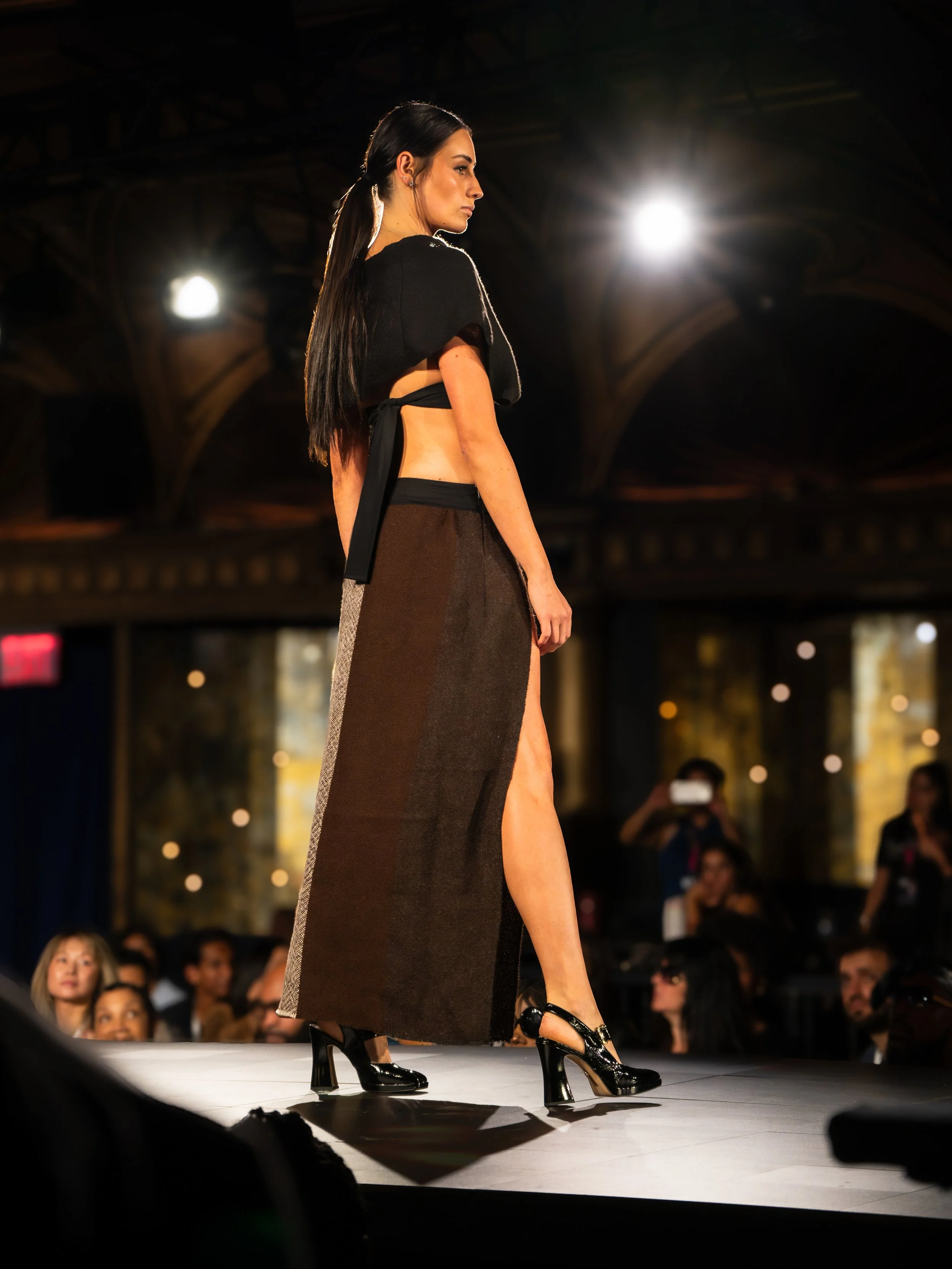 A fashion model walking on runway during fashion show, wearing a black top and long brown skirt with a slit, high heels, with audience and photographers in background.