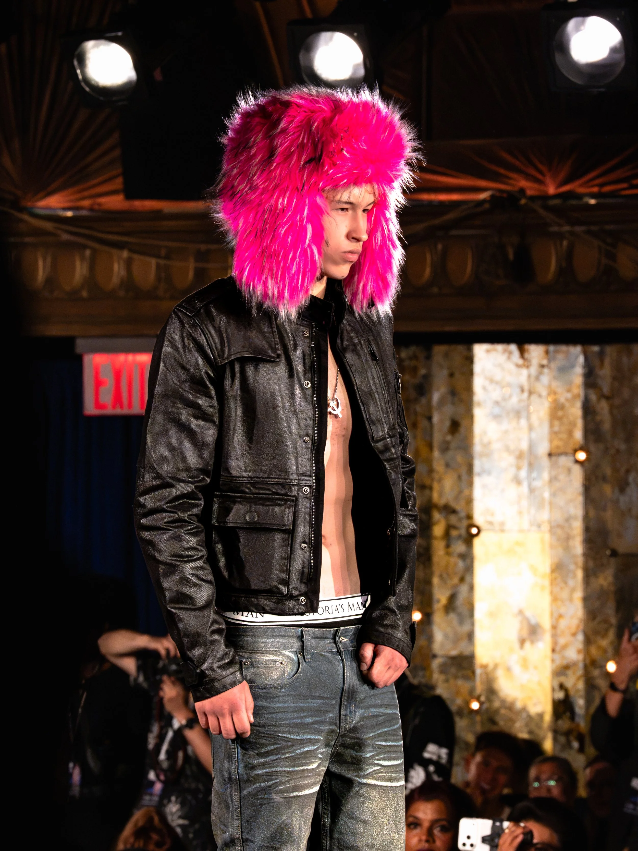A male model on a runway wearing a bright pink furry hat and a black leather jacket during a fashion show.