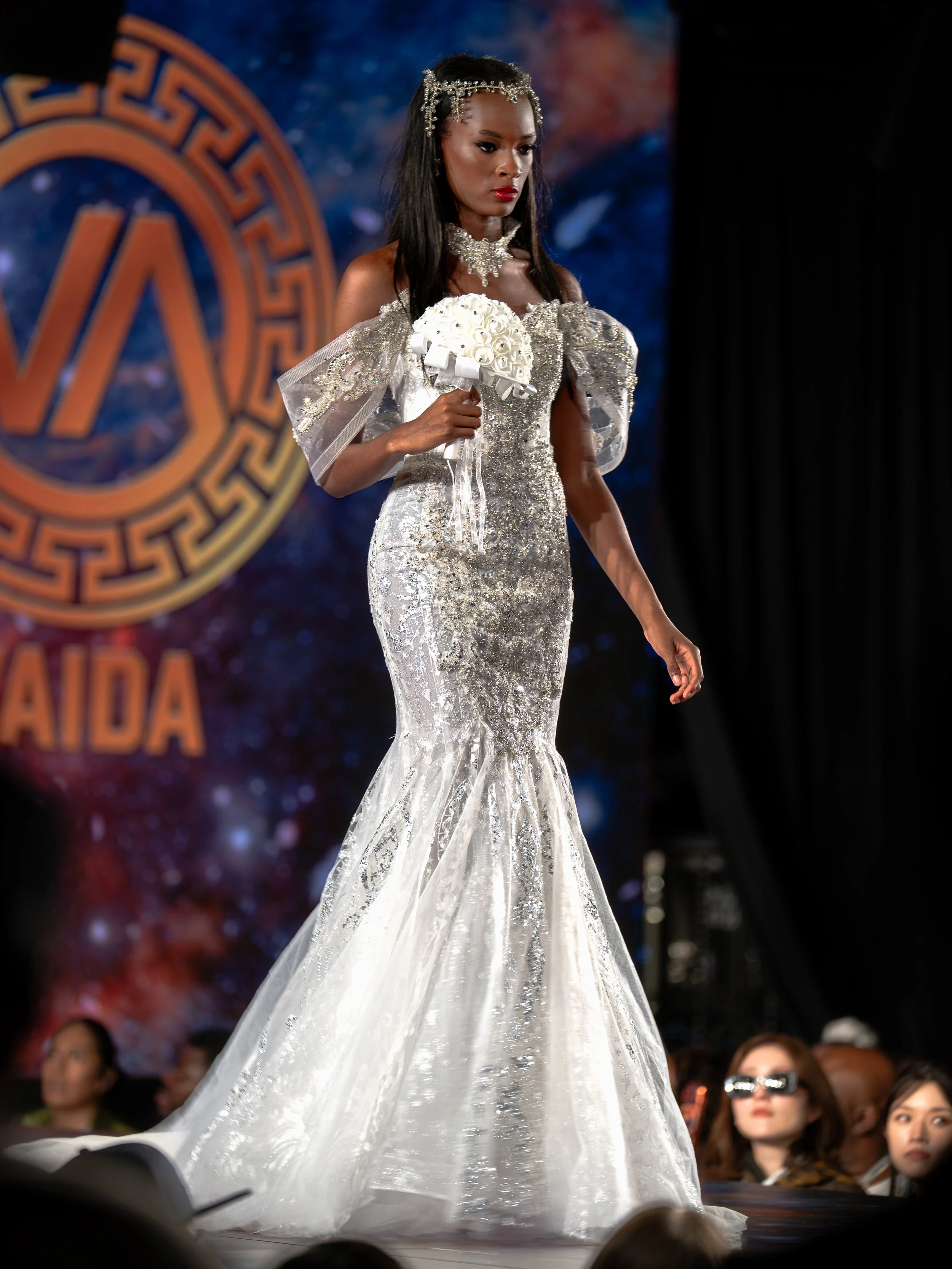 Model walking on stage in a white, elaborate, sequined gown with off-shoulder puffy sleeves, holding a bouquet of white roses, during a fashion show with audience members watching.