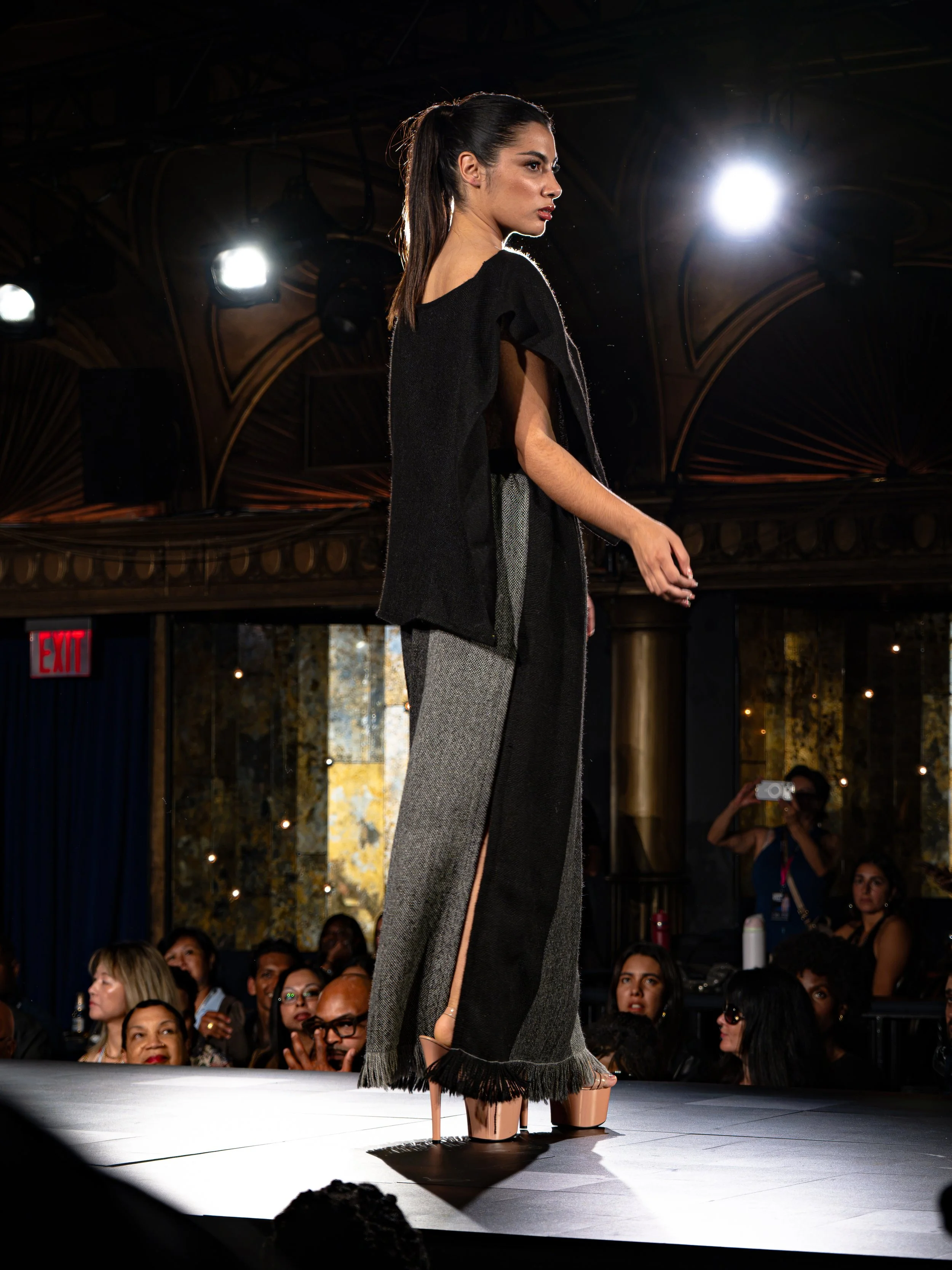 Model walking on runway at fashion show, audience watching, bright lights overhead, ornate architecture in background.