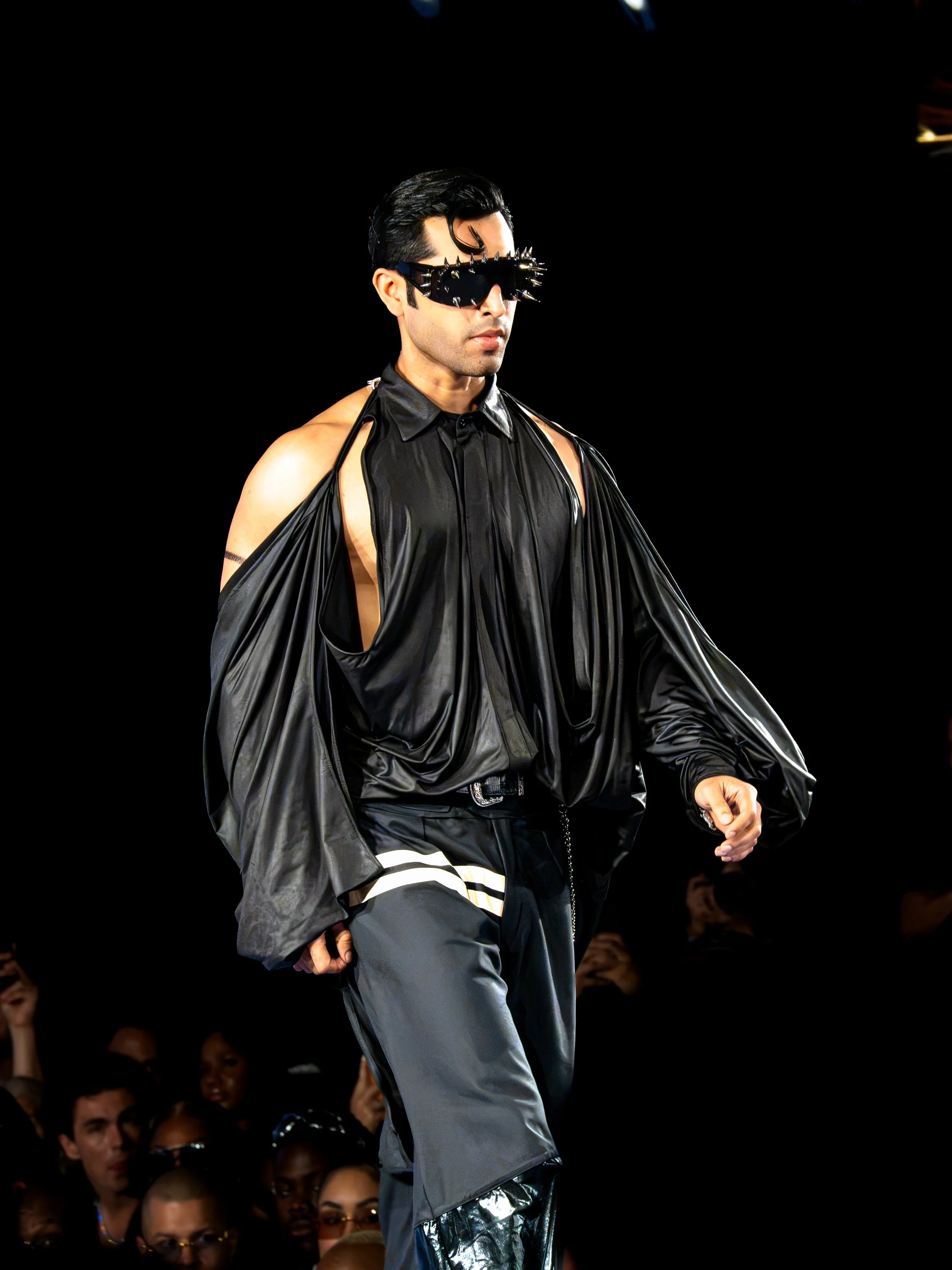 A man walking on a runway during a fashion show, wearing a shiny black cape, gray pants, and accessorized with large, spiky sunglasses.