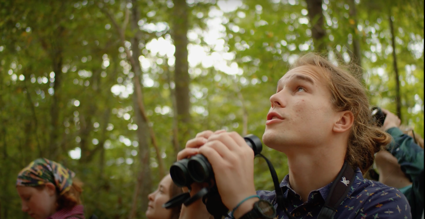 Young man in outdoor clothing holding binoculars and looking upwards in a forest, with other people in the background also engaging in birdwatching.