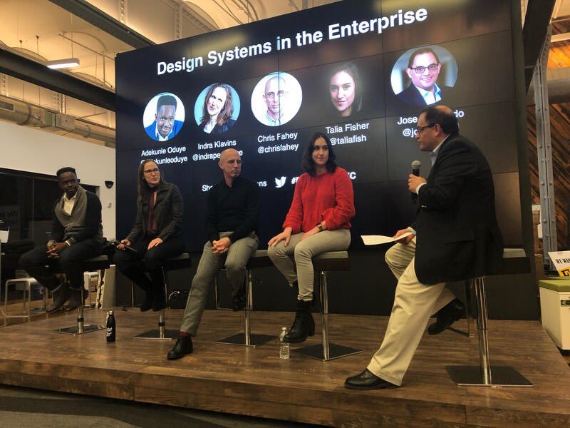 A panel discussion titled "Design Systems in the Enterprise" featuring five speakers sitting on stools on a stage, with a large digital screen behind them displaying their names and social media handles.