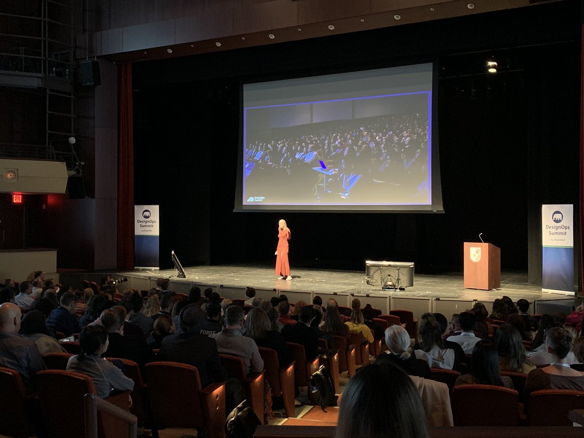 A woman in a red dress giving a presentation on stage at a conference, with a large audience seated in front of her and a big screen displaying the event behind her. There are banners on either side of the stage that read "DesignOps Summit."