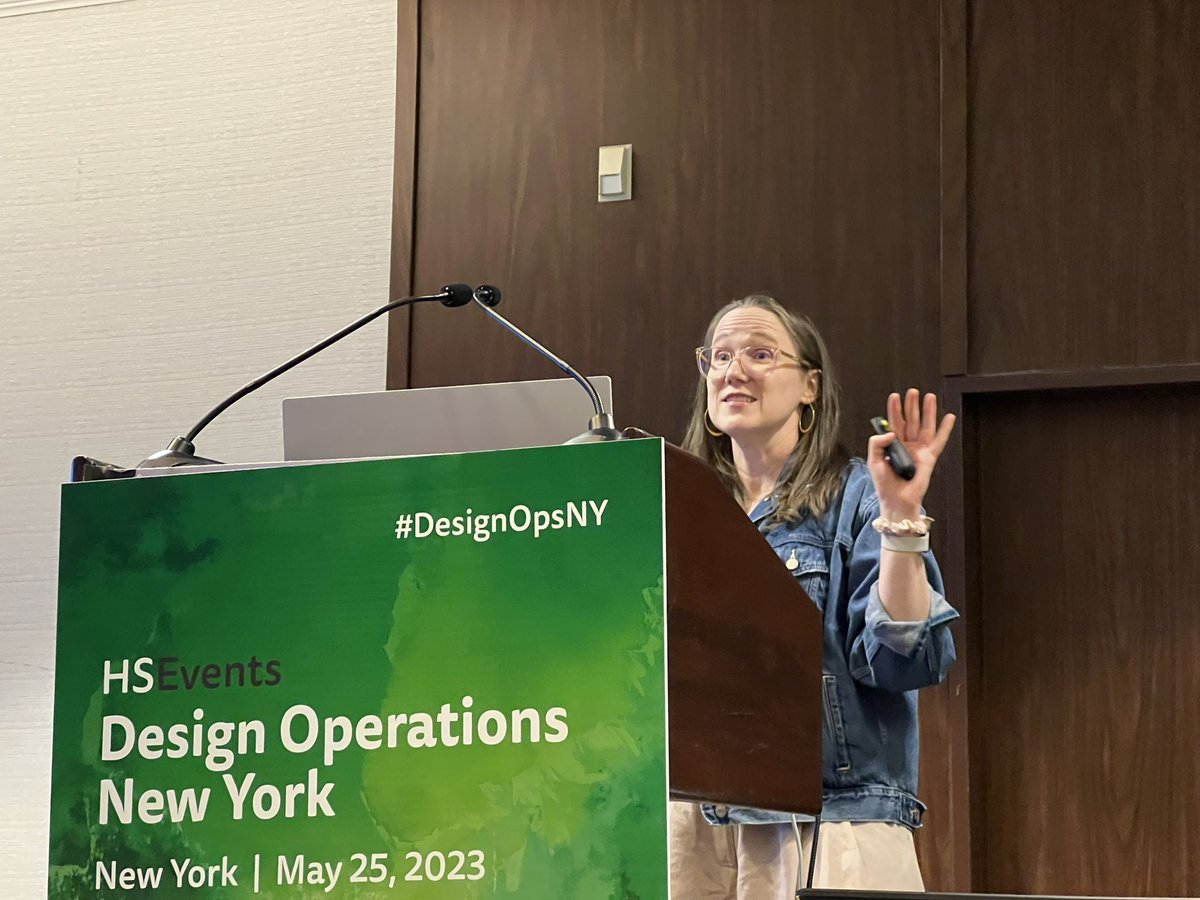 A woman giving a presentation at a conference, standing behind a green podium with the text 'HS Events Design Operations New York May 25, 2023' and a hashtag #DesignOpsNY. She is wearing glasses, a denim jacket, and gold hoop earrings, and is gesturing with her hand.