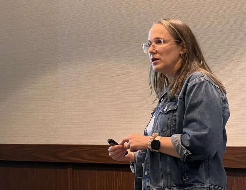 A woman standing indoors, wearing glasses, a denim jacket, and a smartwatch, holding a smartphone, and looking to her left
