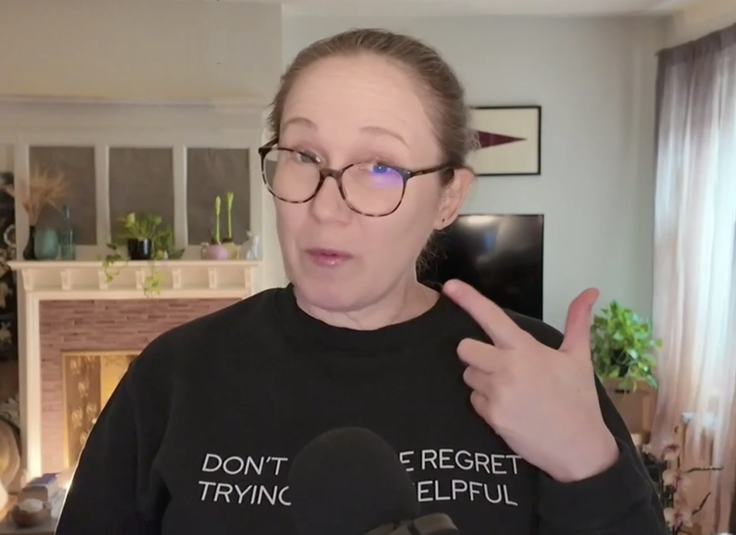 A woman with red hair and glasses pointing to her upper lip, wearing a black shirt with white text, in a living room with a fireplace, plants, and curtains.