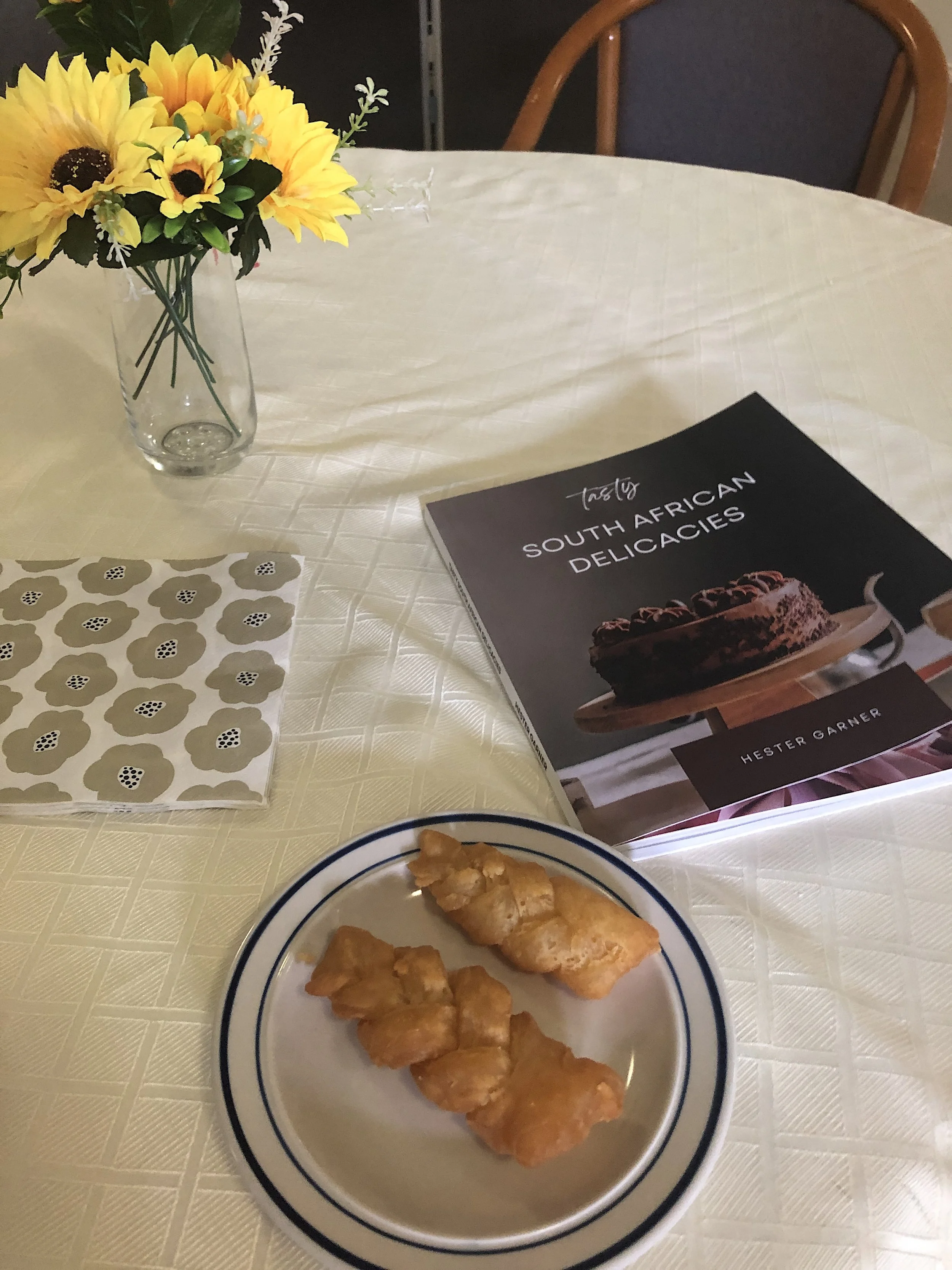 A table with a floral vase, a book titled 'South African Delicacies', a decorative napkin, and a plate of fried snacks.