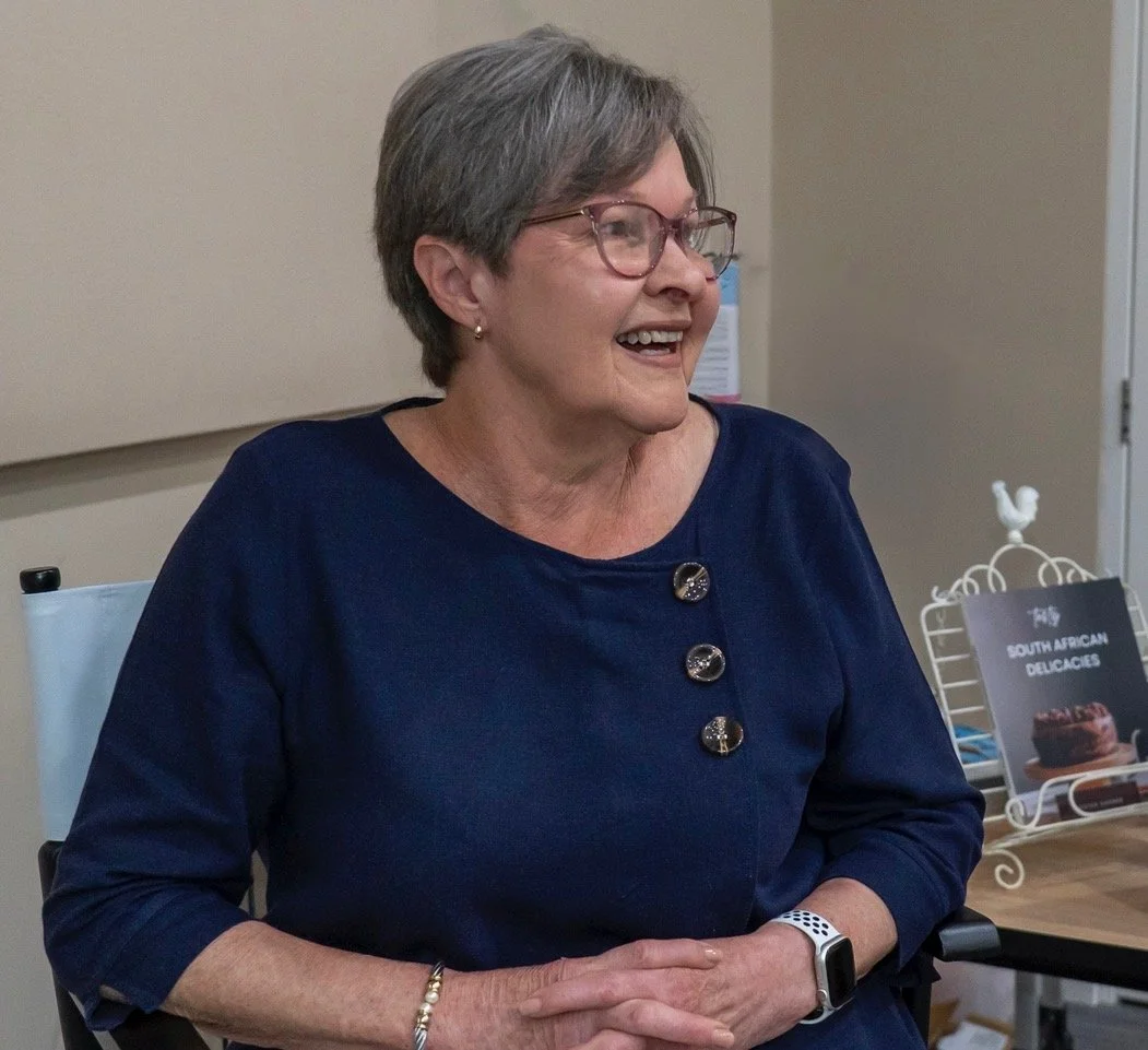 Smiling author, an older woman with short grey hair and glasses, wearing a navy blue top with shiny buttons, sitting at a book display table. A bookstand displays "Tasty South African Recipes".