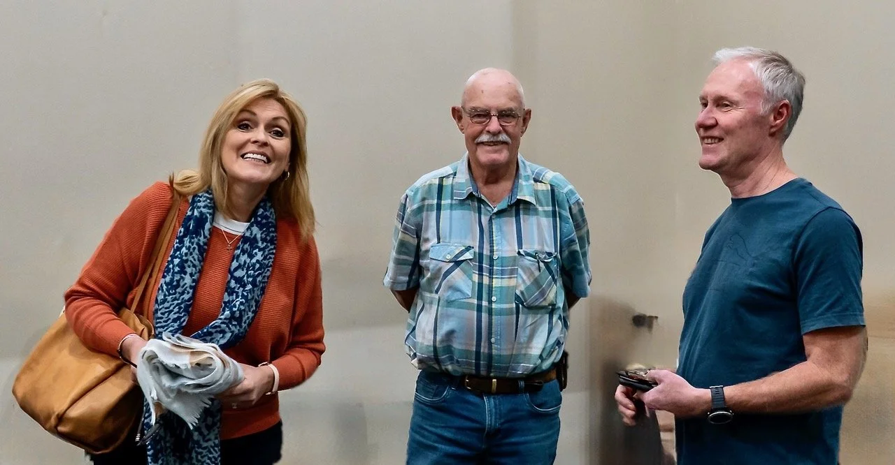 Three people standing together at a book launch, smiling and talking in an indoor setting with a plain wall background.