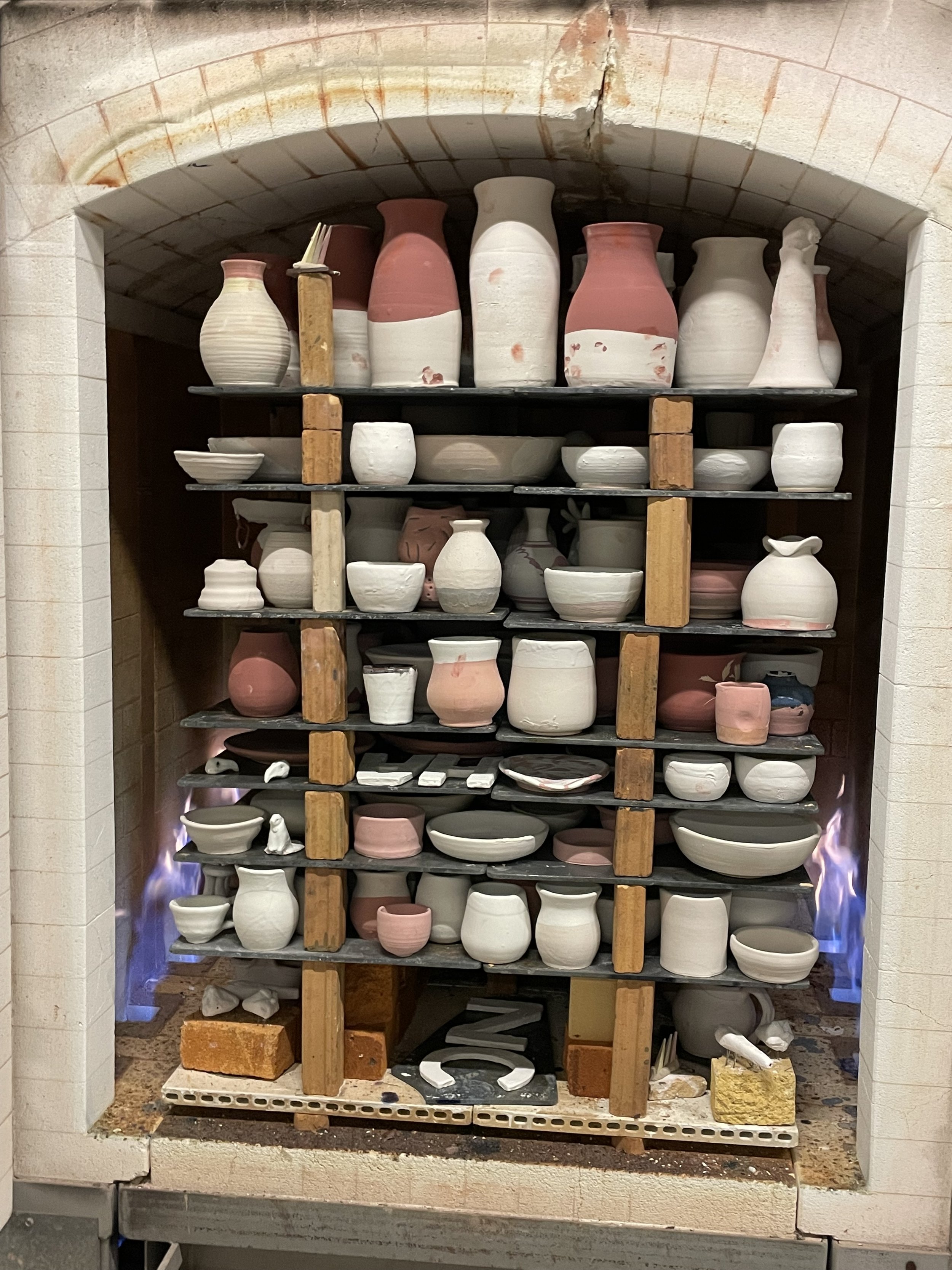 Clay pottery drying in a kiln with multiple shelves holding various unfinished ceramic vases, bowls, and vessels, some pink and others white, with visible bricks and flames at the base.