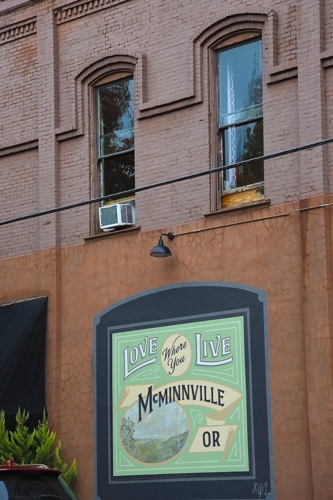 A brick building with two windows, one with an air conditioning unit. A painted mural with the text 'Love Where You Live McMinnville OR' and a landscape circle is on the lower part of the building.