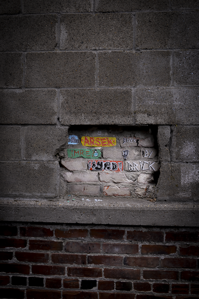 A brick wall with a rectangular hole revealing graffiti on the bricks inside, including colorful tags such as 'ARISEK' and other graffiti art.