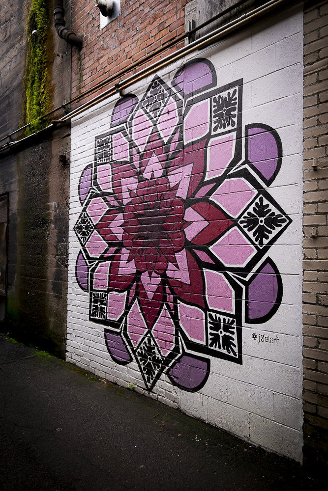 Colorful mural of a pink and purple floral mandala painted on a white brick wall.