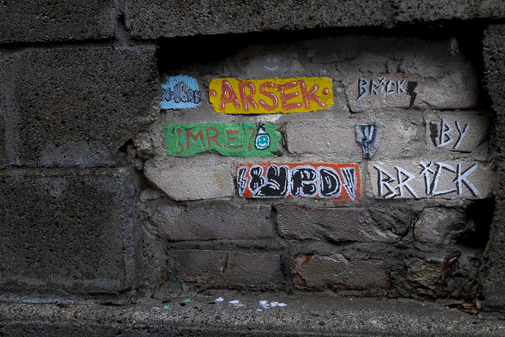 Colorful graffiti on a brick wall with words and symbols, including 'ARЗEK,' 'BRICK,' and 'SMREZ,' in various colors and styles.