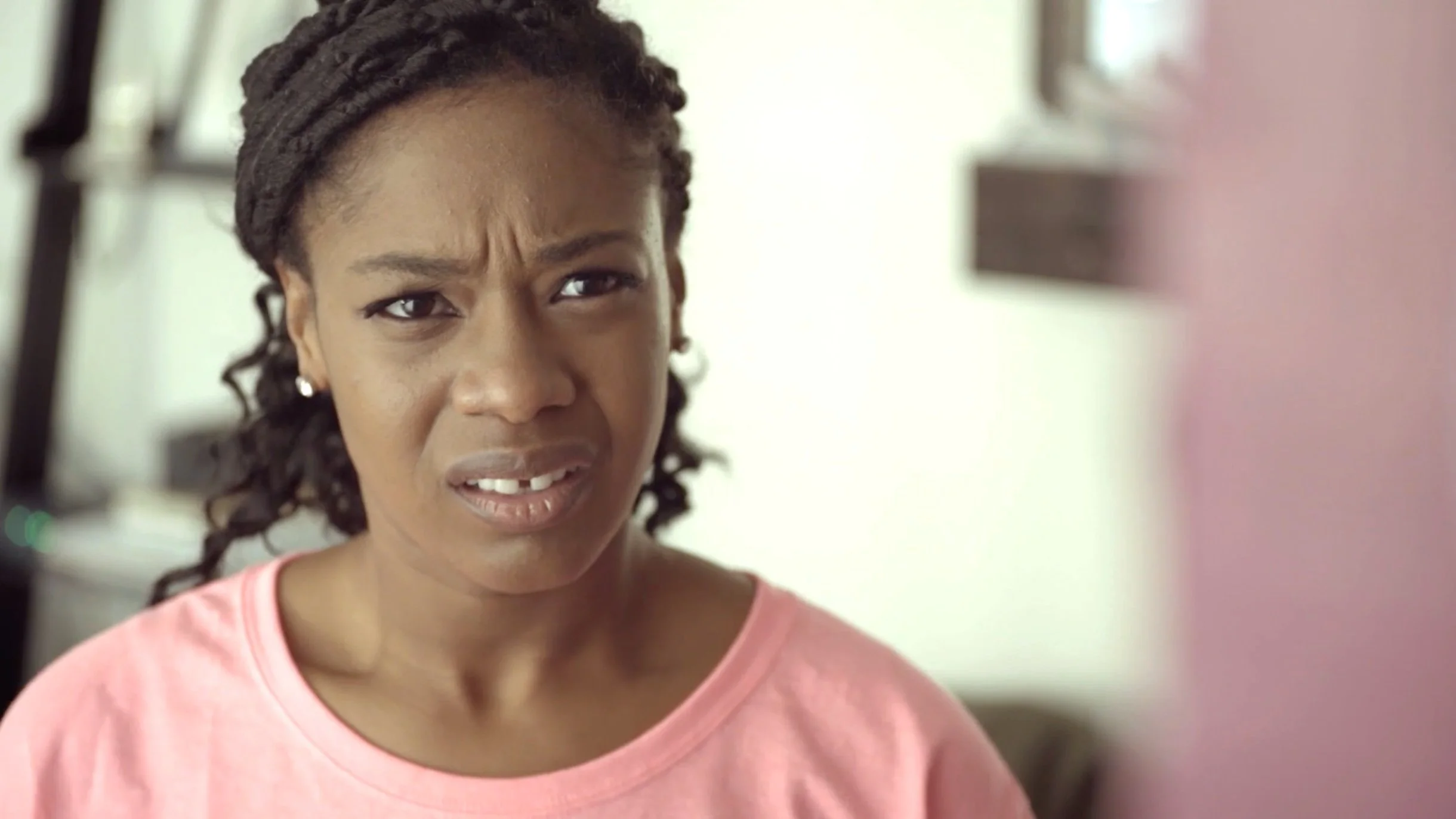 Close-up of a woman with a confused expression, wearing a pink shirt and earrings, indoors.