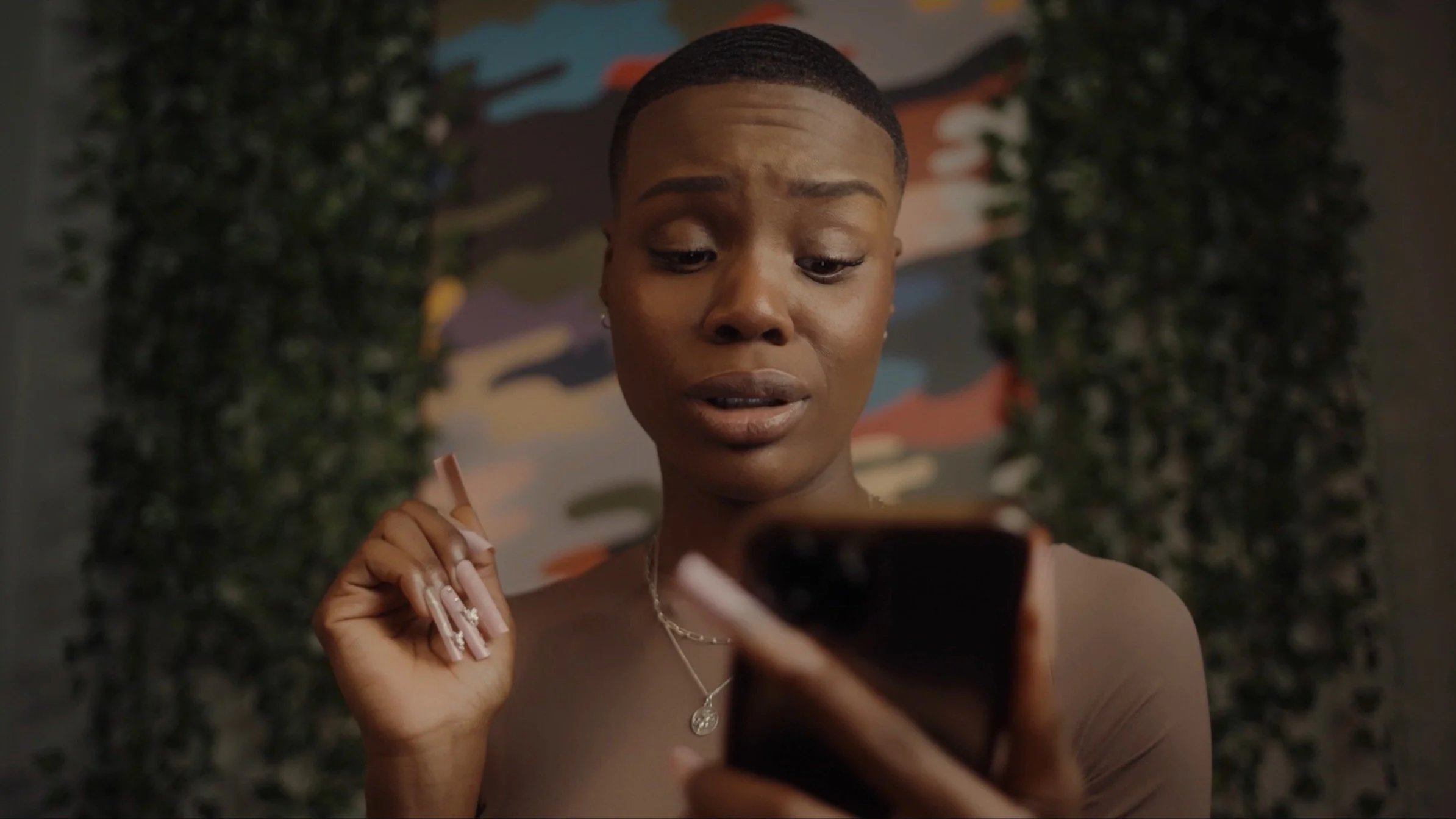 Person with short hair looking at a smartphone, holding it with manicured fingers, in front of a vibrant abstract background and green foliage.