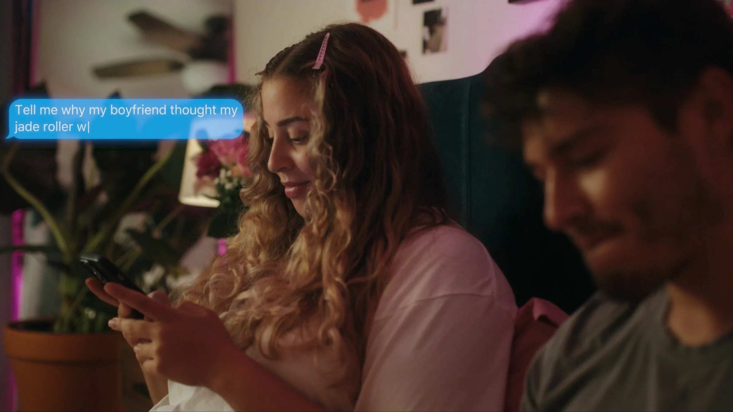A woman with curly hair and a hair clip is smiling and looking at her phone while sitting next to a man. A blue text bubble appears next to her, reading, "Tell me why my boyfriend thought my jade roller w[...]." There are plants and decorations in th