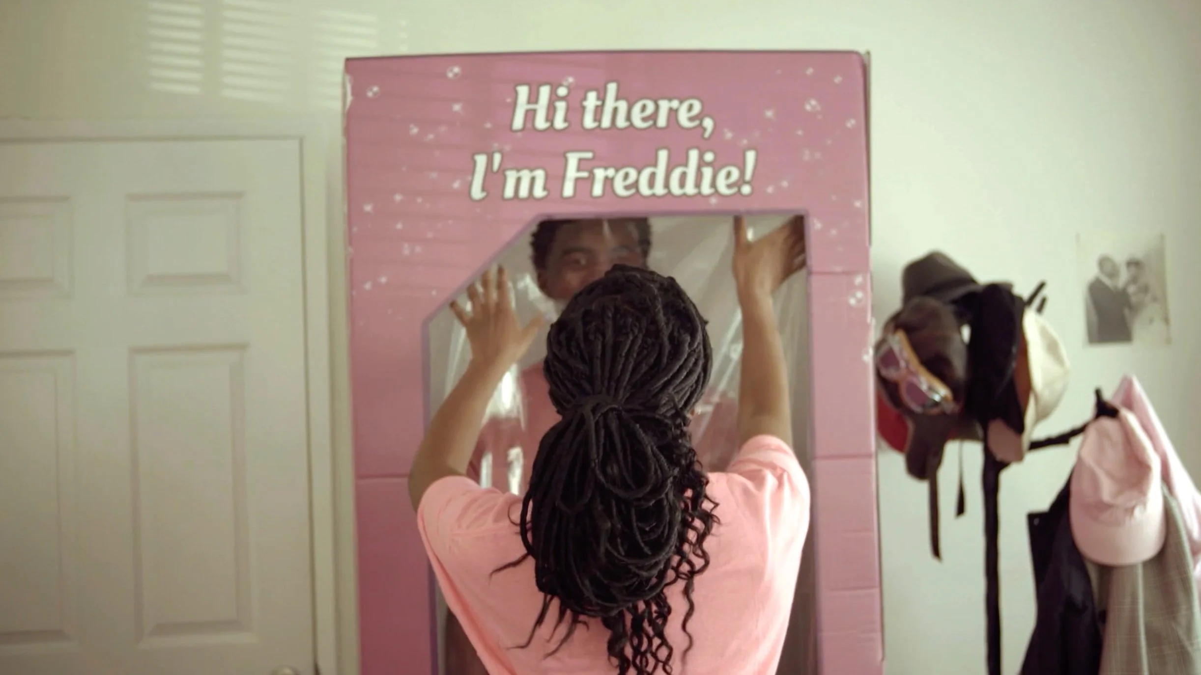 Person putting a cardboard box with "Hi there, I'm Freddie!" text over someone else, in a room with hats and clothing hanging nearby.