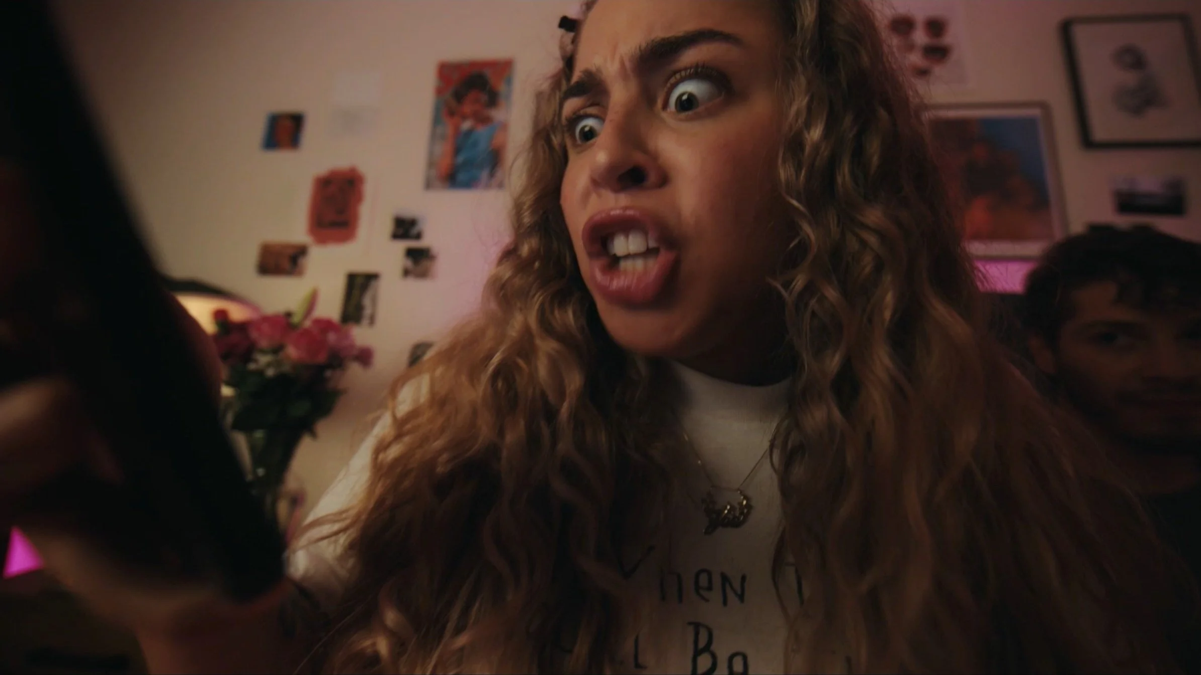 A woman with long curly hair showing an angry expression, holding a smartphone. The background features a room with various photos and decorations on the wall, including flowers and another person partially visible.