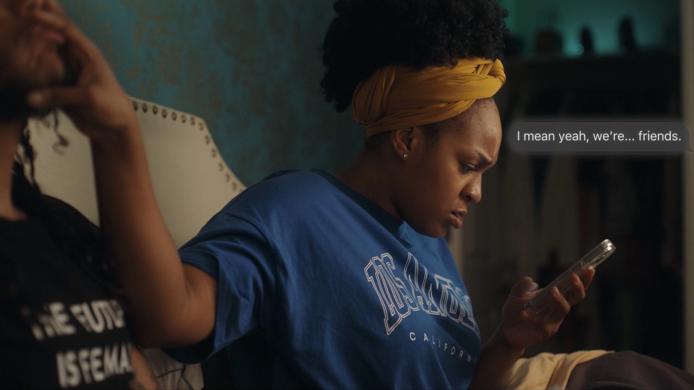 A woman in a blue Los Angeles t-shirt and yellow headwrap sitting on a bed, looking at her phone. Text beside her reads, "I mean yeah, we're... friends." Another person with long hair and a black t-shirt is partially visible.
