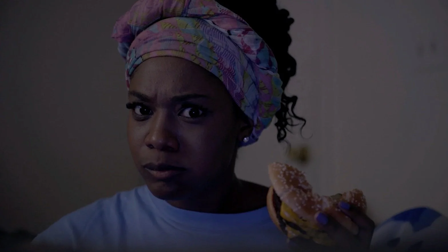 A person with a serious expression, wearing a colorful headscarf, holding a cheeseburger.