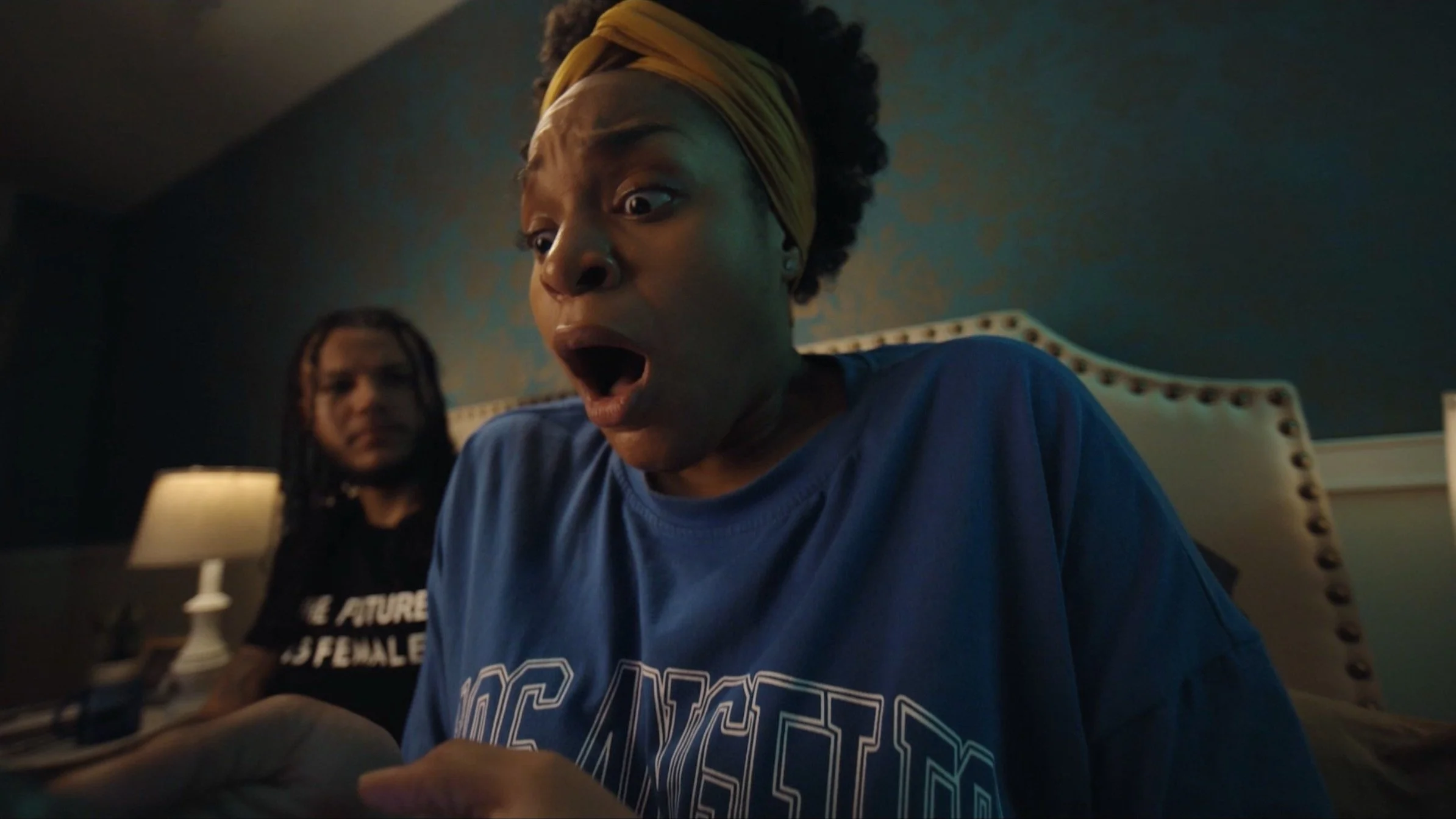 Two people in a dimly lit room, one in the foreground wearing a blue shirt with "Los Angeles" on it, looking surprised, with a person in the background wearing a black shirt with partial text visible.