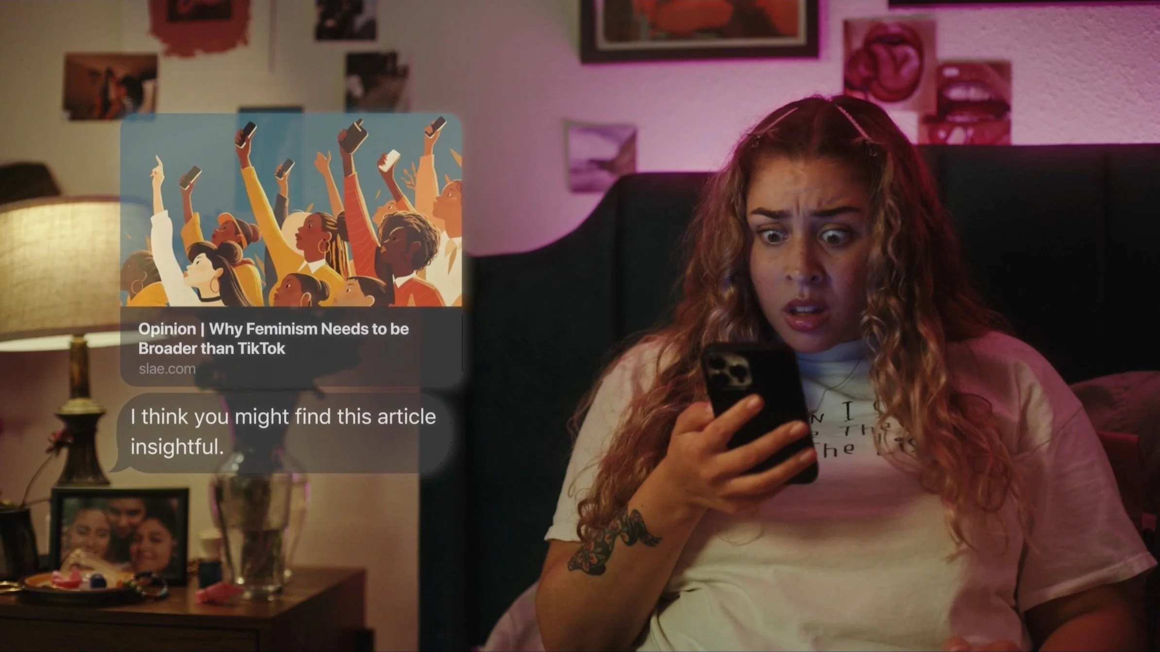 A surprised woman with curly hair looks at her phone while sitting on a bed. A message with an article suggestion reads: "Opinion | Why Feminism Needs to be Broader than TikTok" alongside an illustration of diverse people holding smartphones. The room has a lamp, pictures on the wall, and framed photos. The atmosphere is dimly lit with a pink hue.