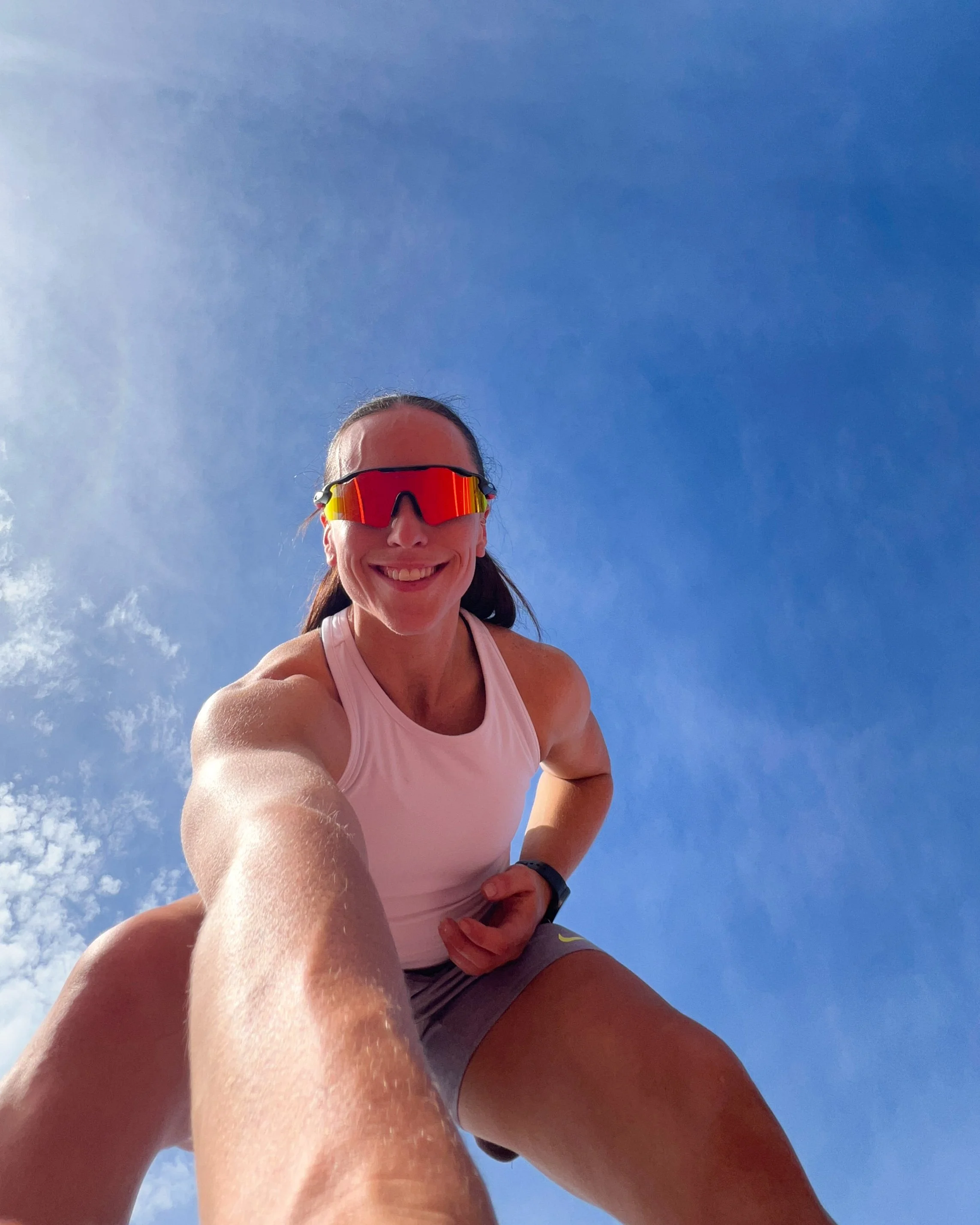A woman smiling wearing sunglasses, a white tank top, and shorts, taken from a low angle with a clear blue sky in the background.