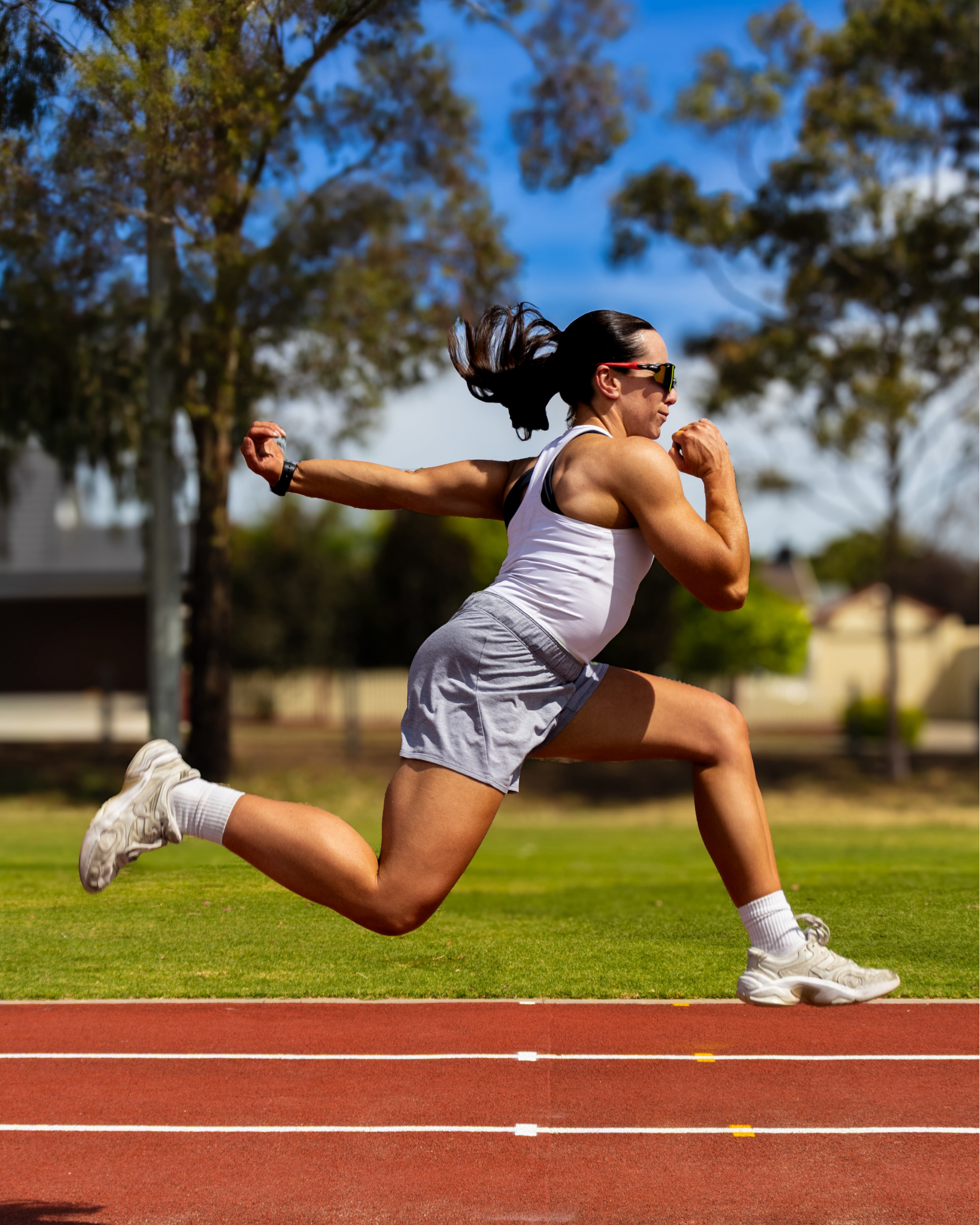 A woman running on a track, captured mid-stride with her hair flowing and wearing sunglasses, athletic tank top, shorts, and running shoes, outdoors on a sunny day.