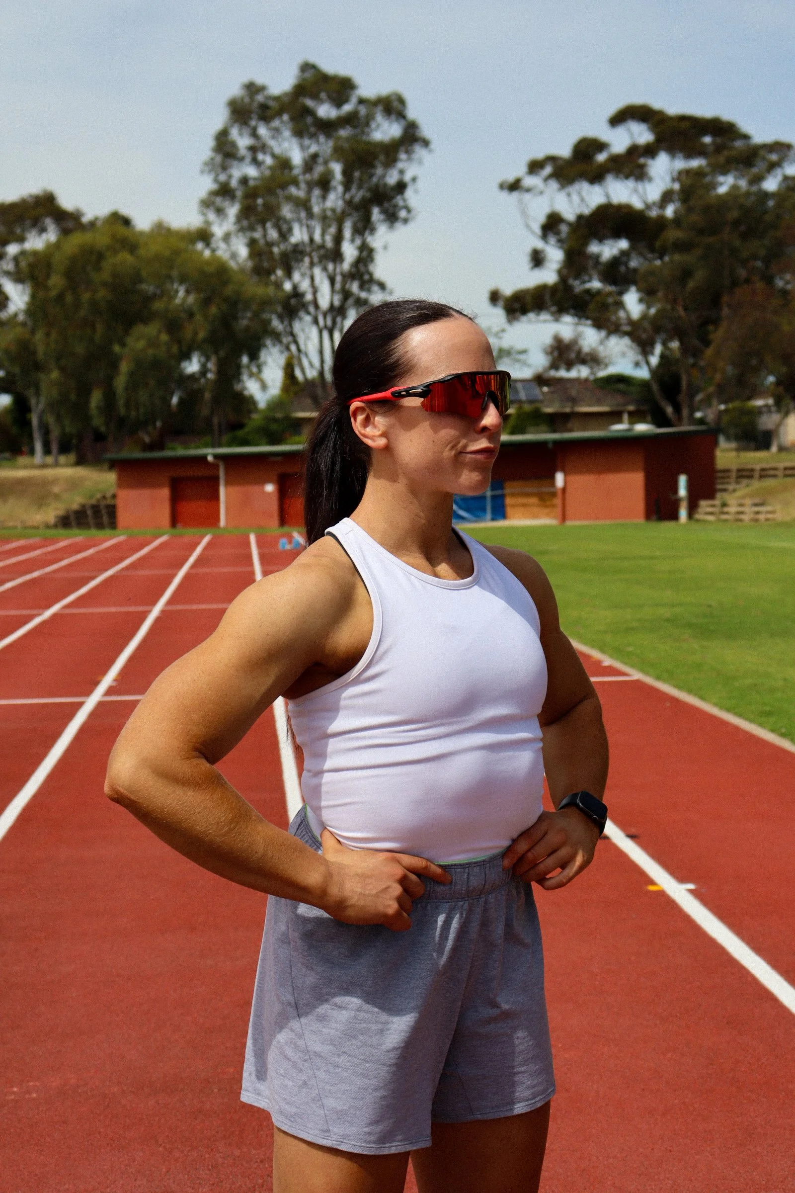 female athlete standing on a running track wearing sunglasses, a white sleeveless top, and gray shorts, with her hands on her hips, in a stadium like outdoor setting.
