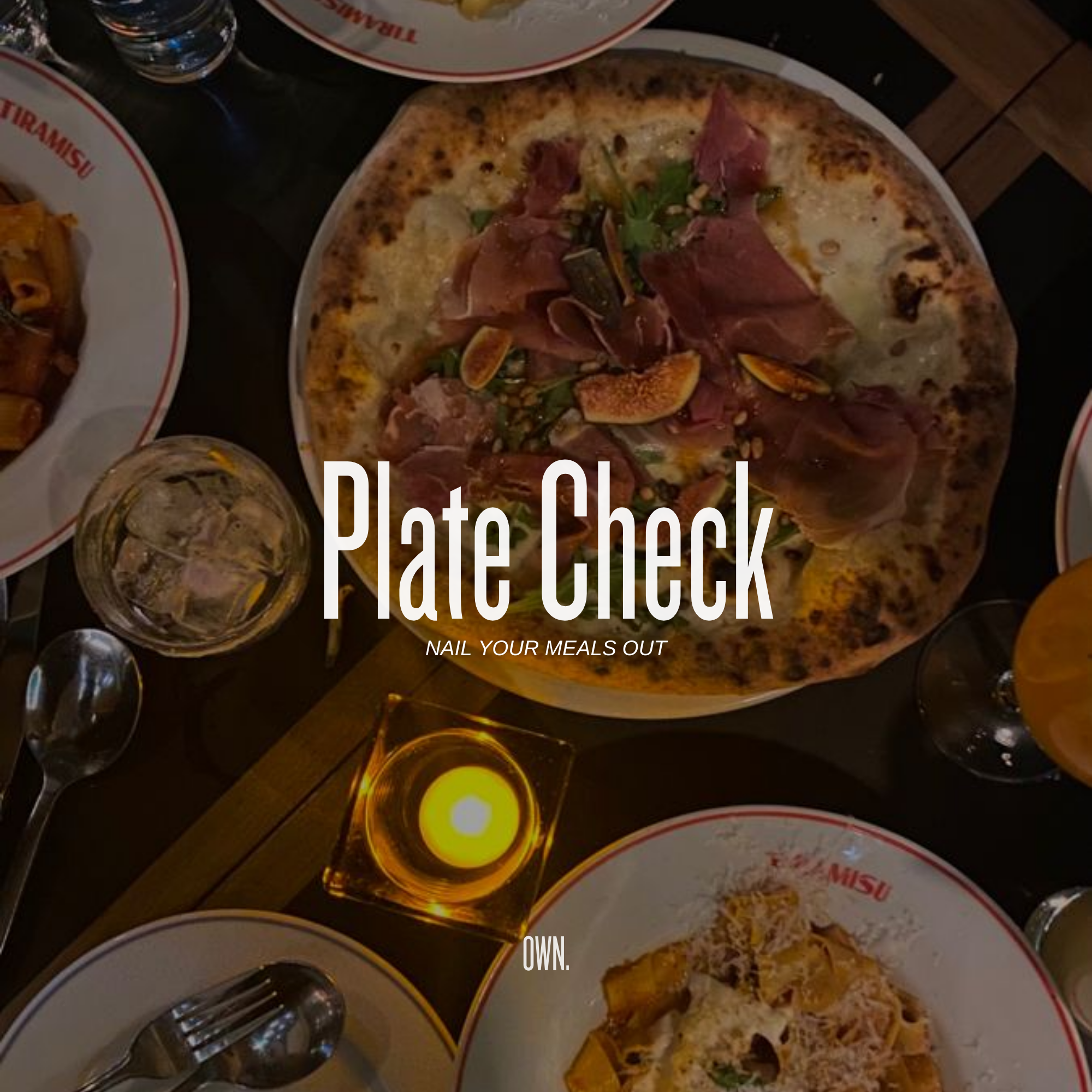 A table set with various dishes, including a large pizza with meats and greens, a glass of white wine, a candle, and plates of pasta, covered with a dark tablecloth.