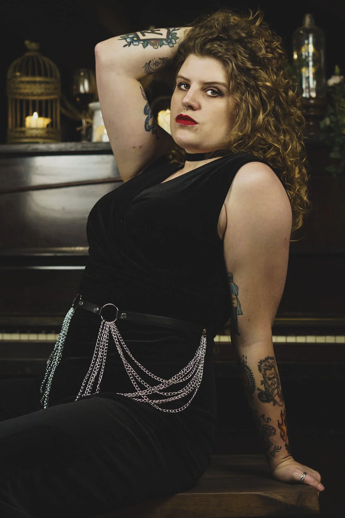 Portrait of woman in black velvet gothic jumpsuit with chains on belt sat in front of piano one hand on head in sexy pose