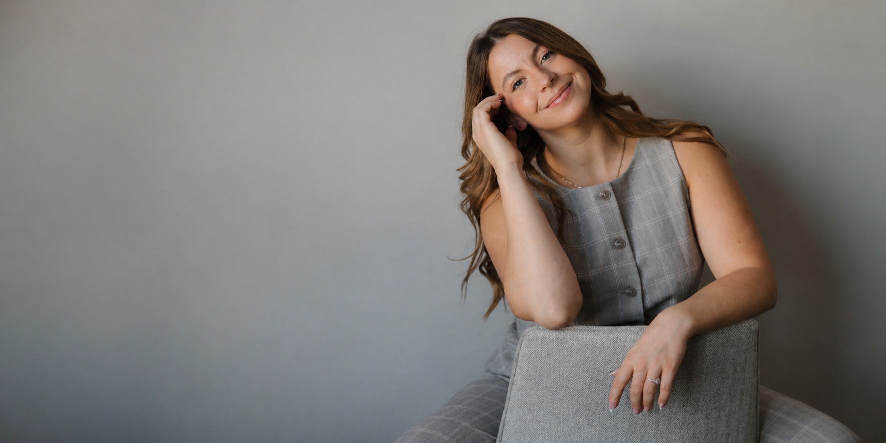 A woman with long wavy brown hair smiling and resting her head on her hand, sitting on a chair against a plain gray background.