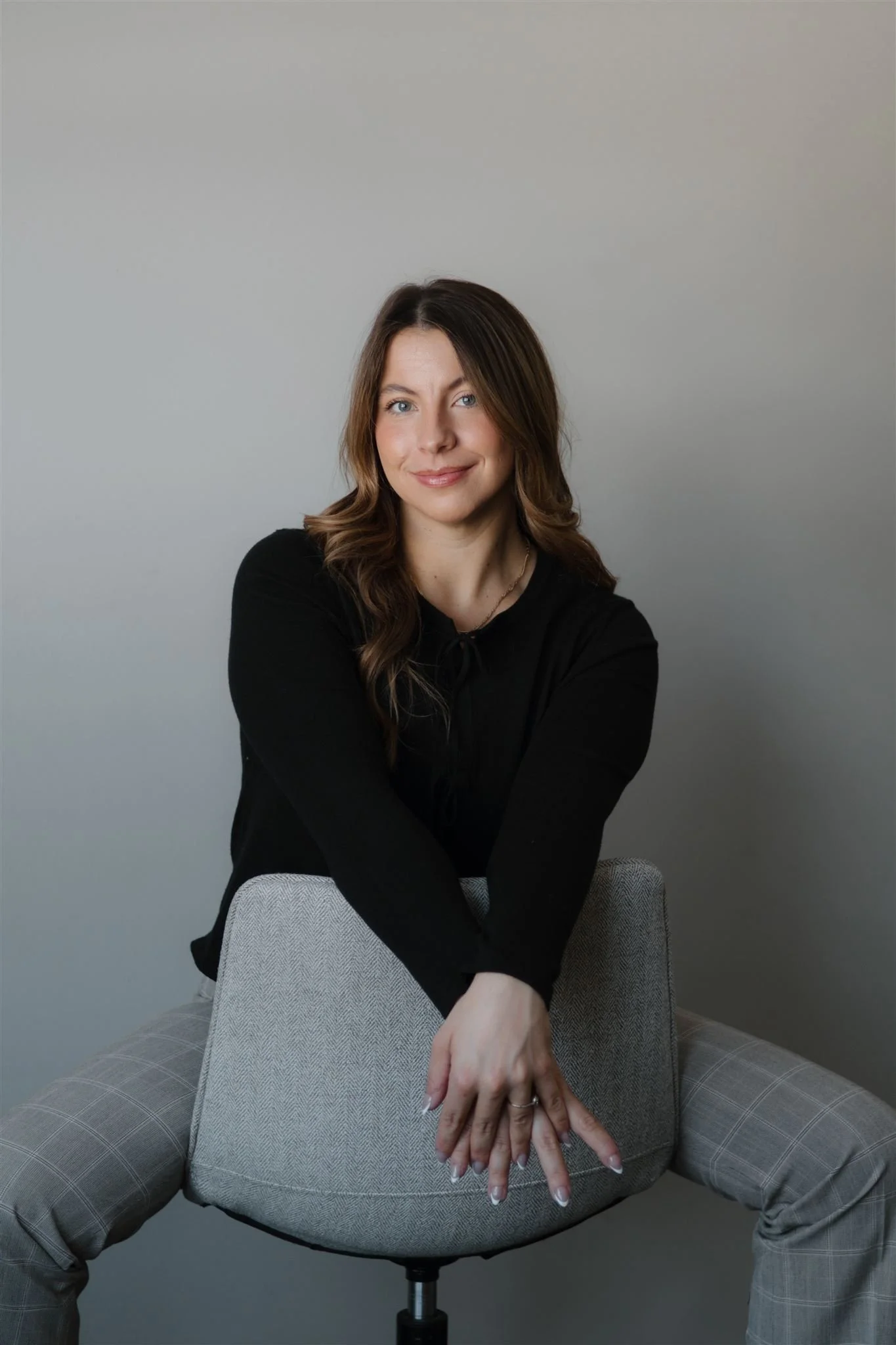 A woman with brown wavy hair wearing a black top and gray plaid pants is sitting on a gray fabric chair, smiling and looking at the camera, with her hands resting on her knees against a plain gray wall background.
