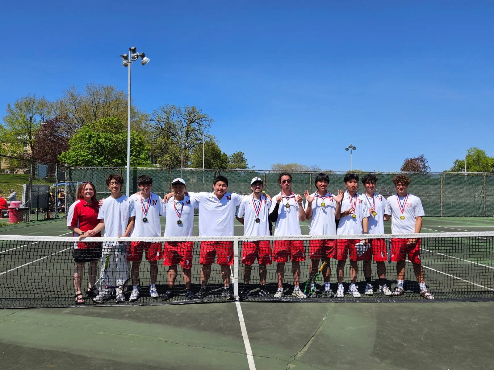 A tennis team of twelve people, including a coach, standing on a tennis court after a match with medals around their necks, under a clear blue sky.