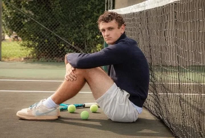 Young man sitting on a tennis court with four tennis balls and a tennis racket, leaning against the net.
