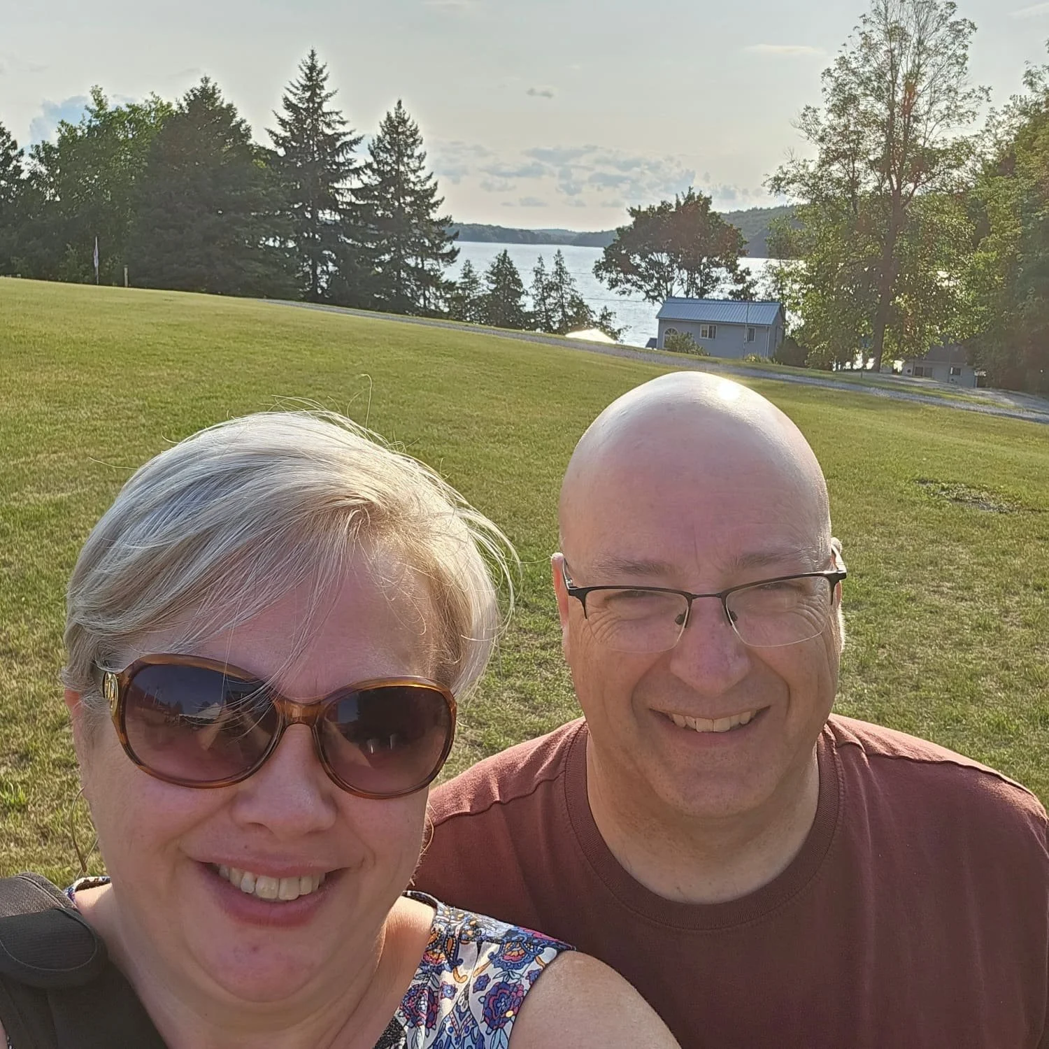 Smiling couple taking a selfie outdoors with a grassy field, trees, and a lake in the background.